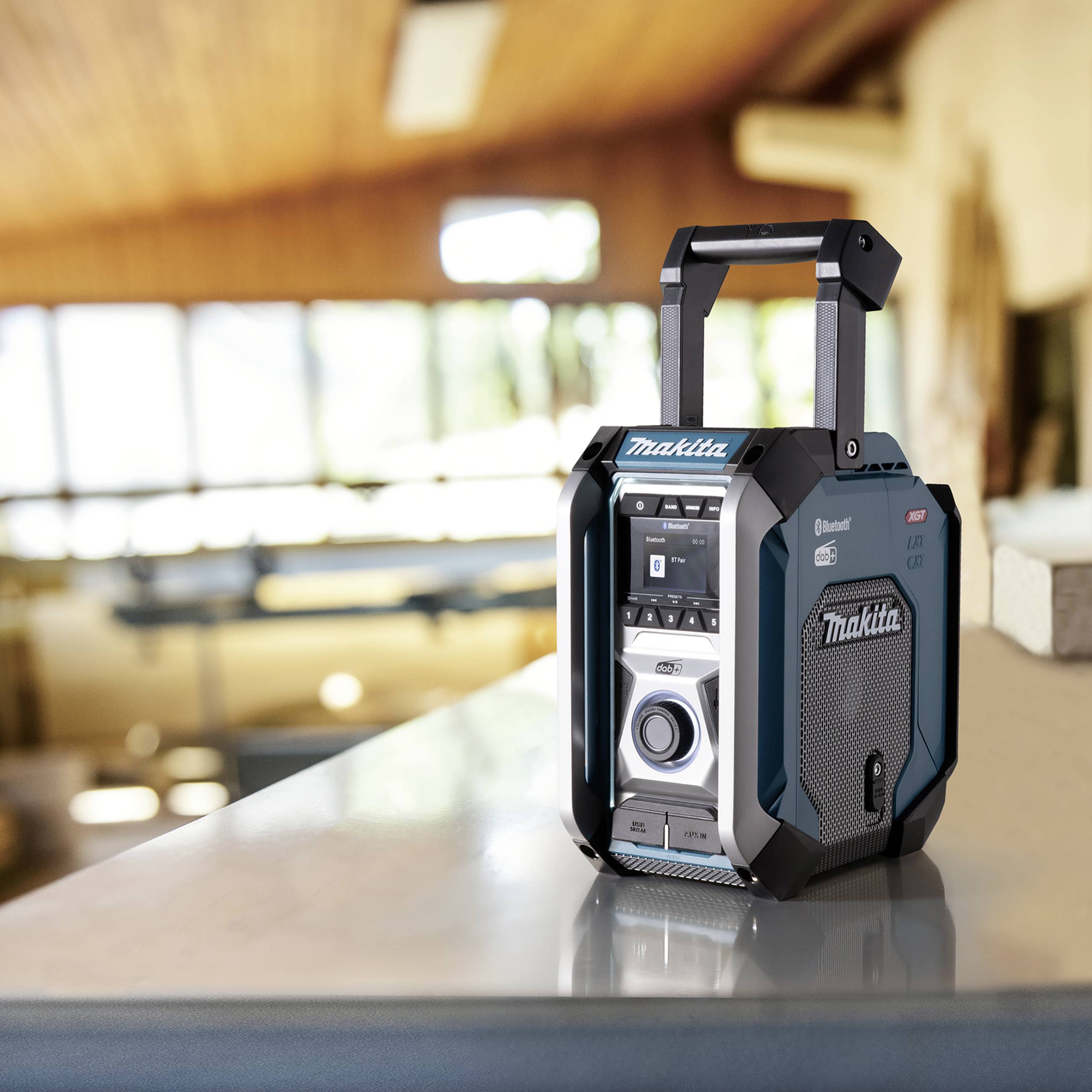 Een draagbare radio met Bluetooth-functie van het merk Makita staat op een tafel in een lichte ruimte met houten elementen en ramen.