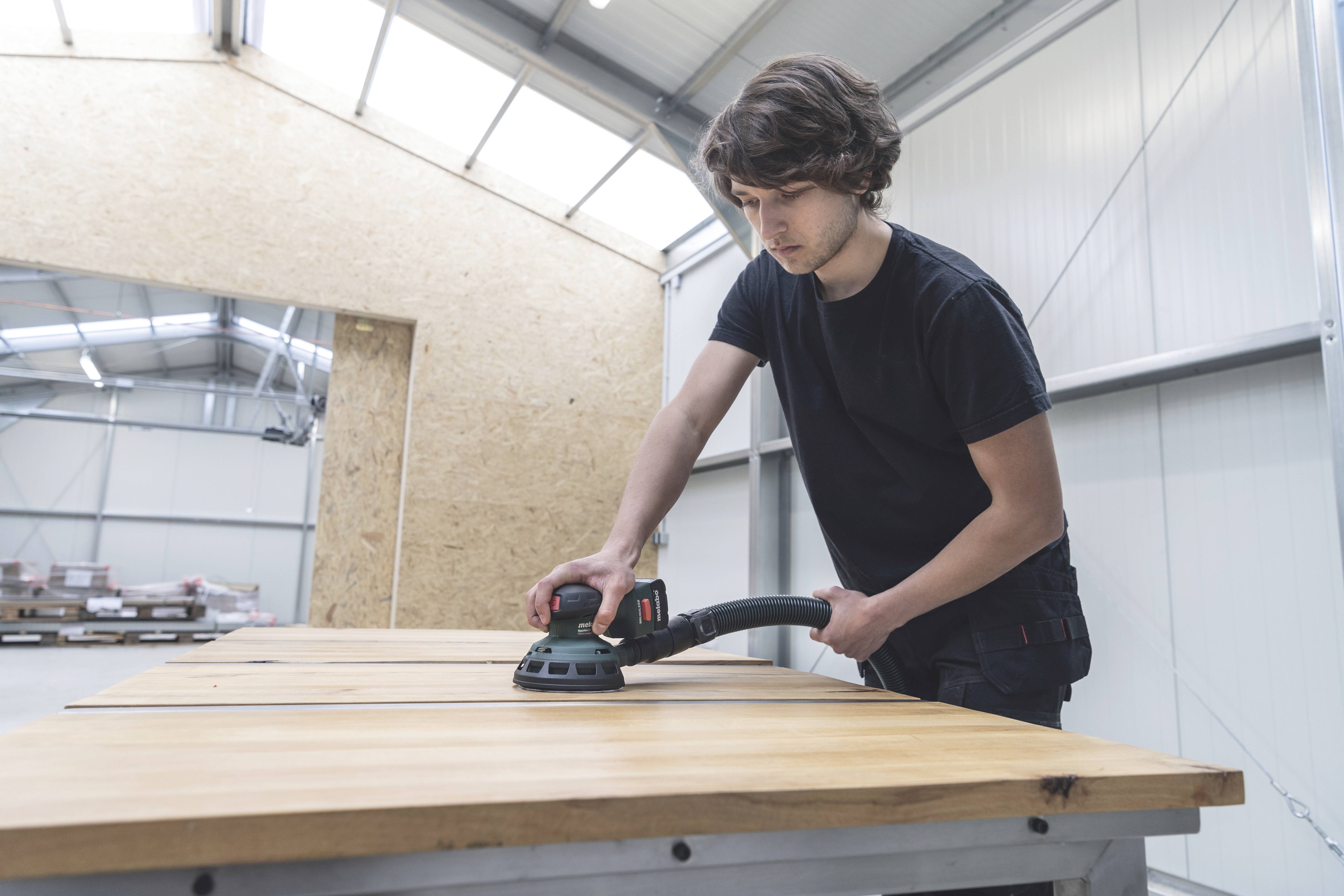 Een man slijpt een grote houten plaat met een elektrisch slijpapparaat in een werkplaats.