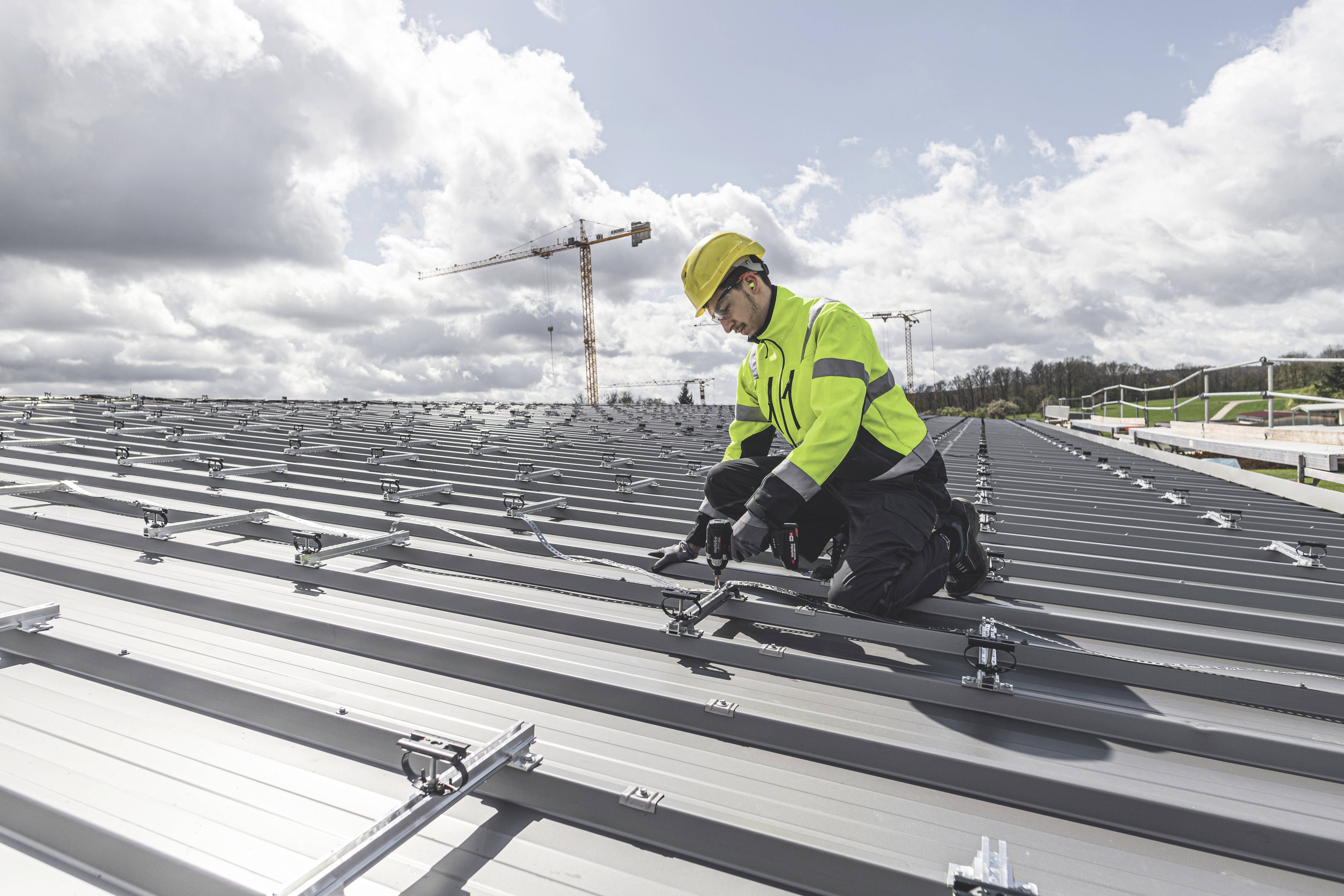 Een arbeider monteert zonnepanelen op een metalen dak, gekleed in beschermende werkkleding, helm en handschoenen. Op de achtergrond een hijskraan en wolken.