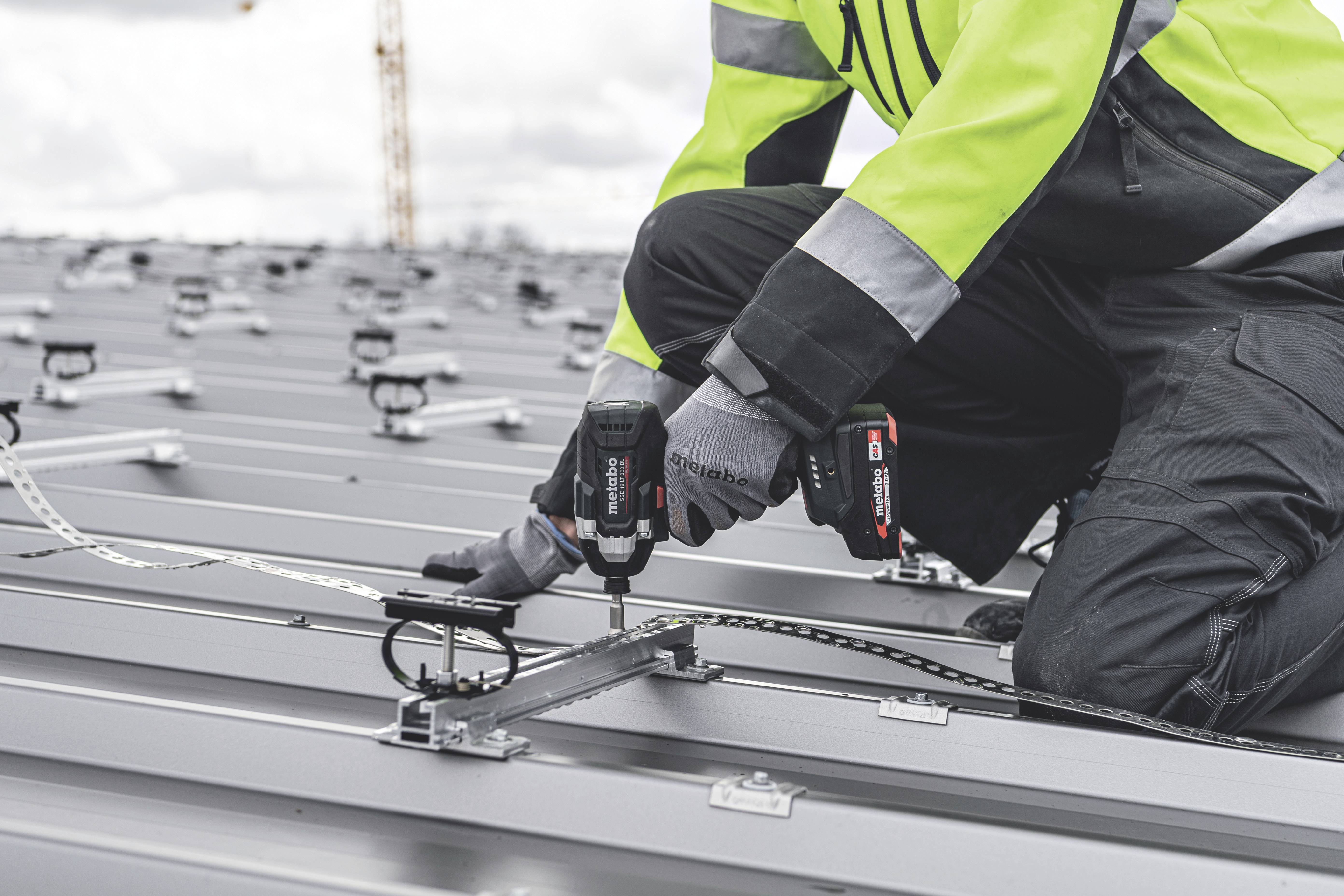 Een persoon in werkkleding installeert zonnepanelen op een metalen dak met behulp van een elektrische boormachine.
