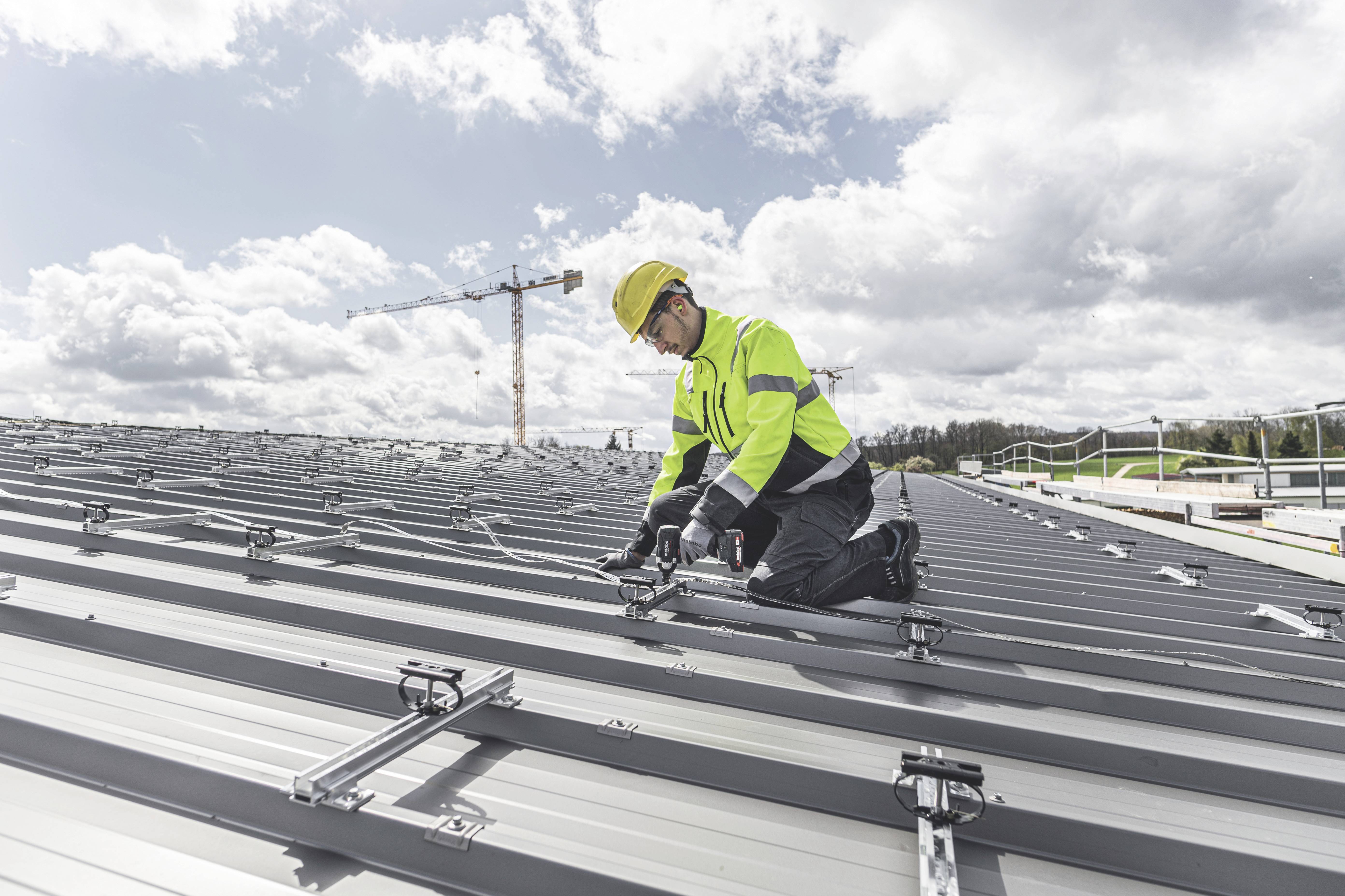 Een persoon in beschermende kleding installeert zonnepanelen op een metalen dak, met op de achtergrond een hijskraan.