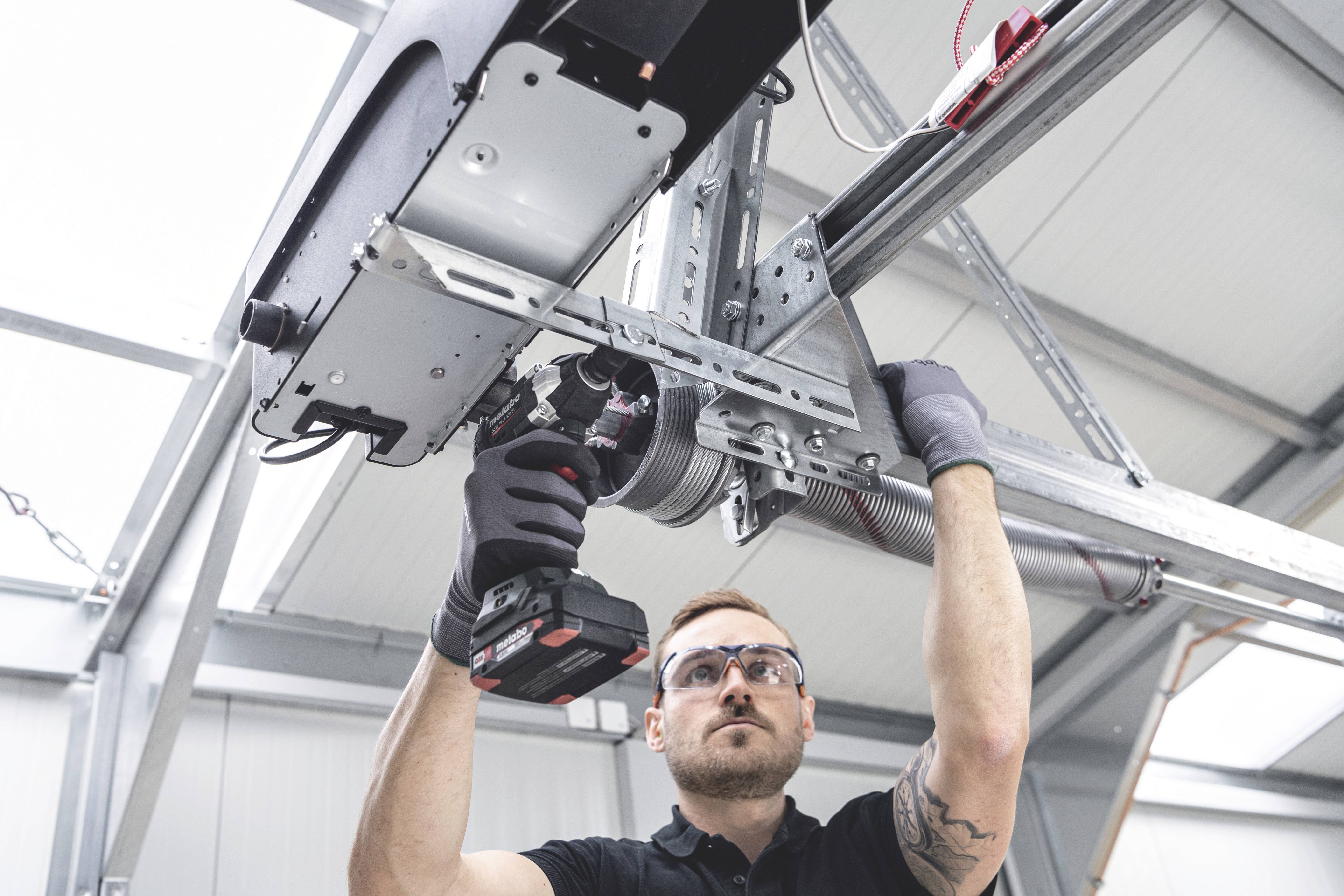 Een man repareert een garagedeur. Hij draagt een veiligheidsbril en gebruikt een elektrisch gereedschap, in een werkplaatsomgeving.