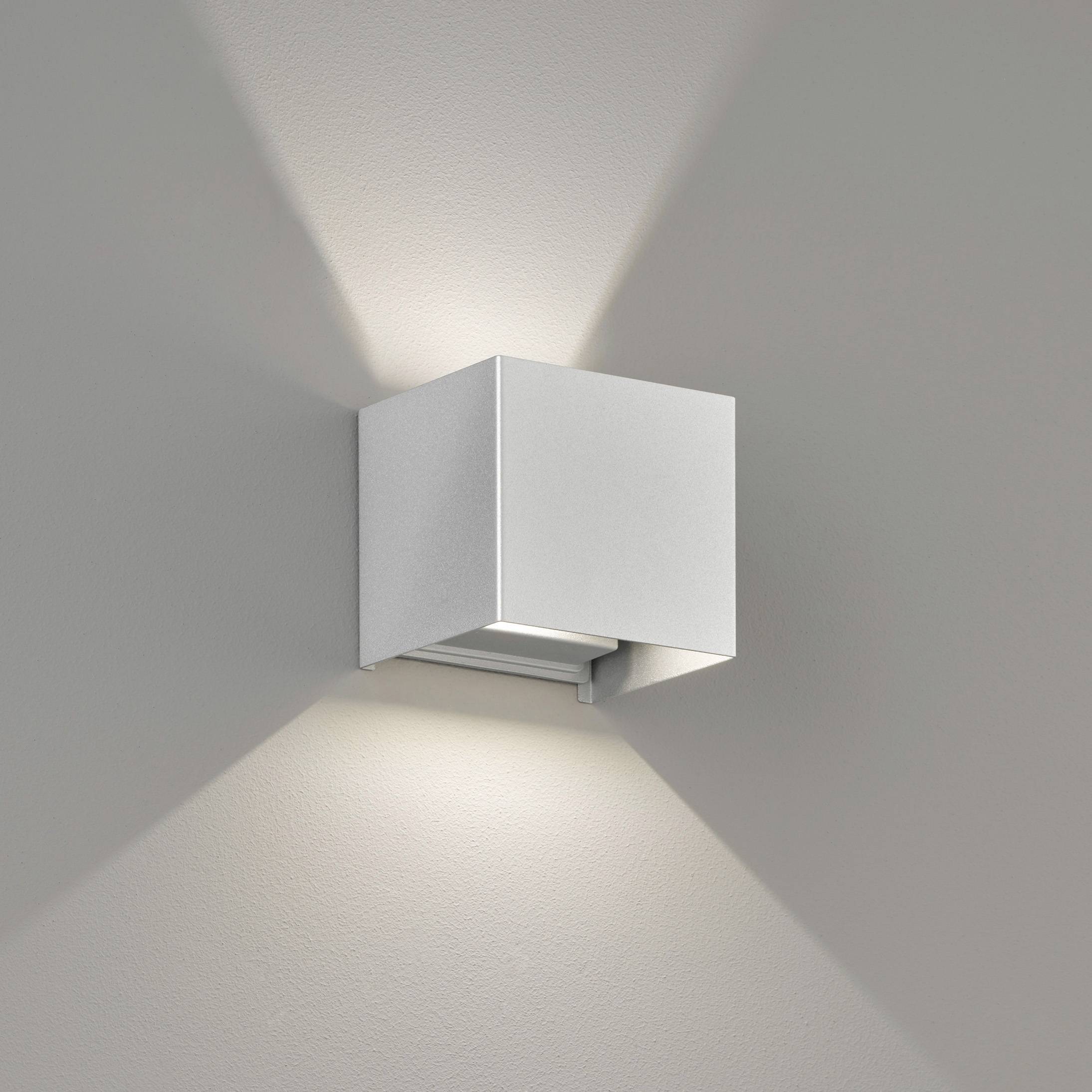 Wandlamp van metaal met hoekig ontwerp, die zowel naar boven als naar beneden schijnt, gemonteerd op een grijze muur.