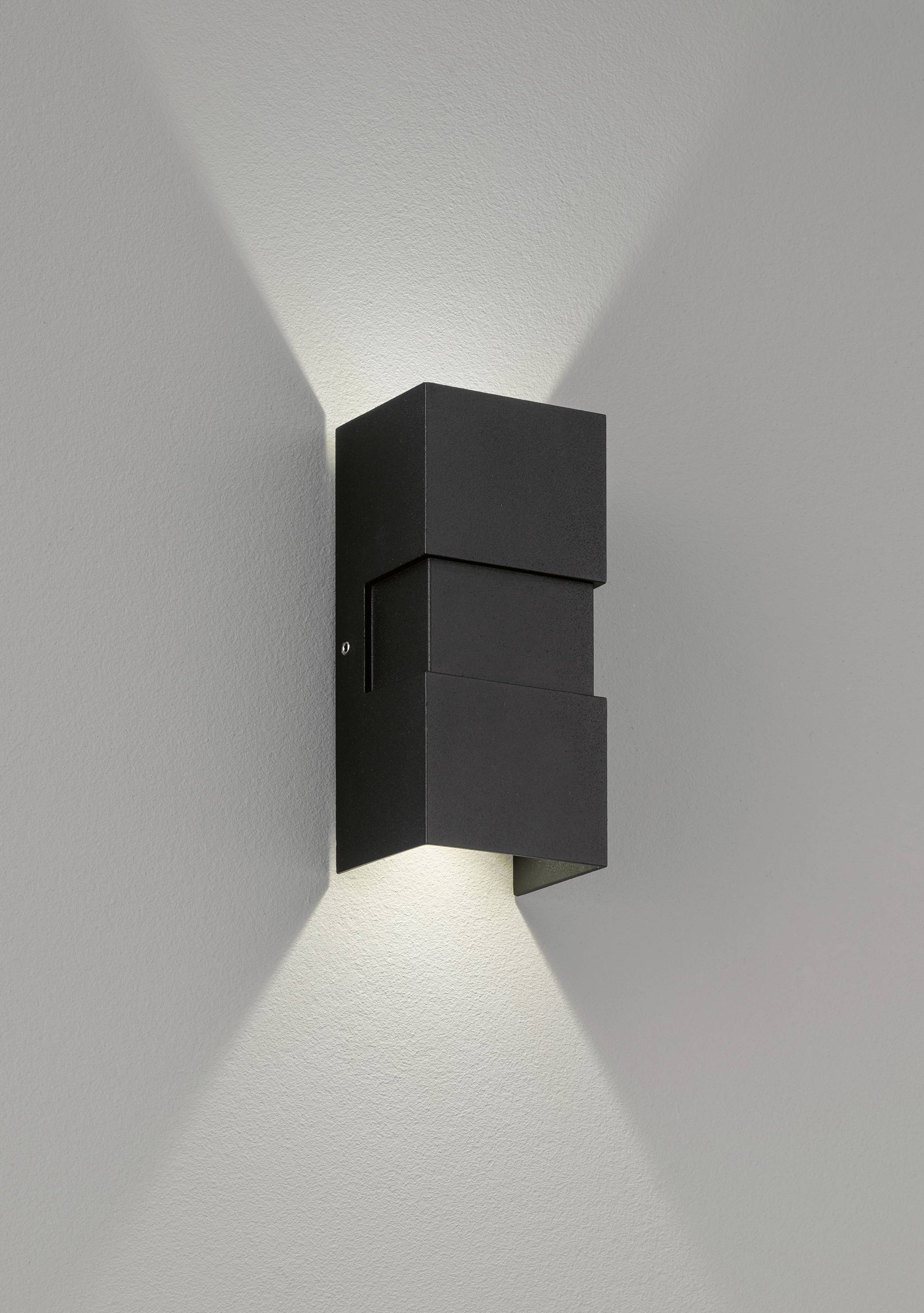 Zwarte wandlamp straalt licht naar boven en beneden tegen een lichte muur, creëert geometrische lichtpatronen.