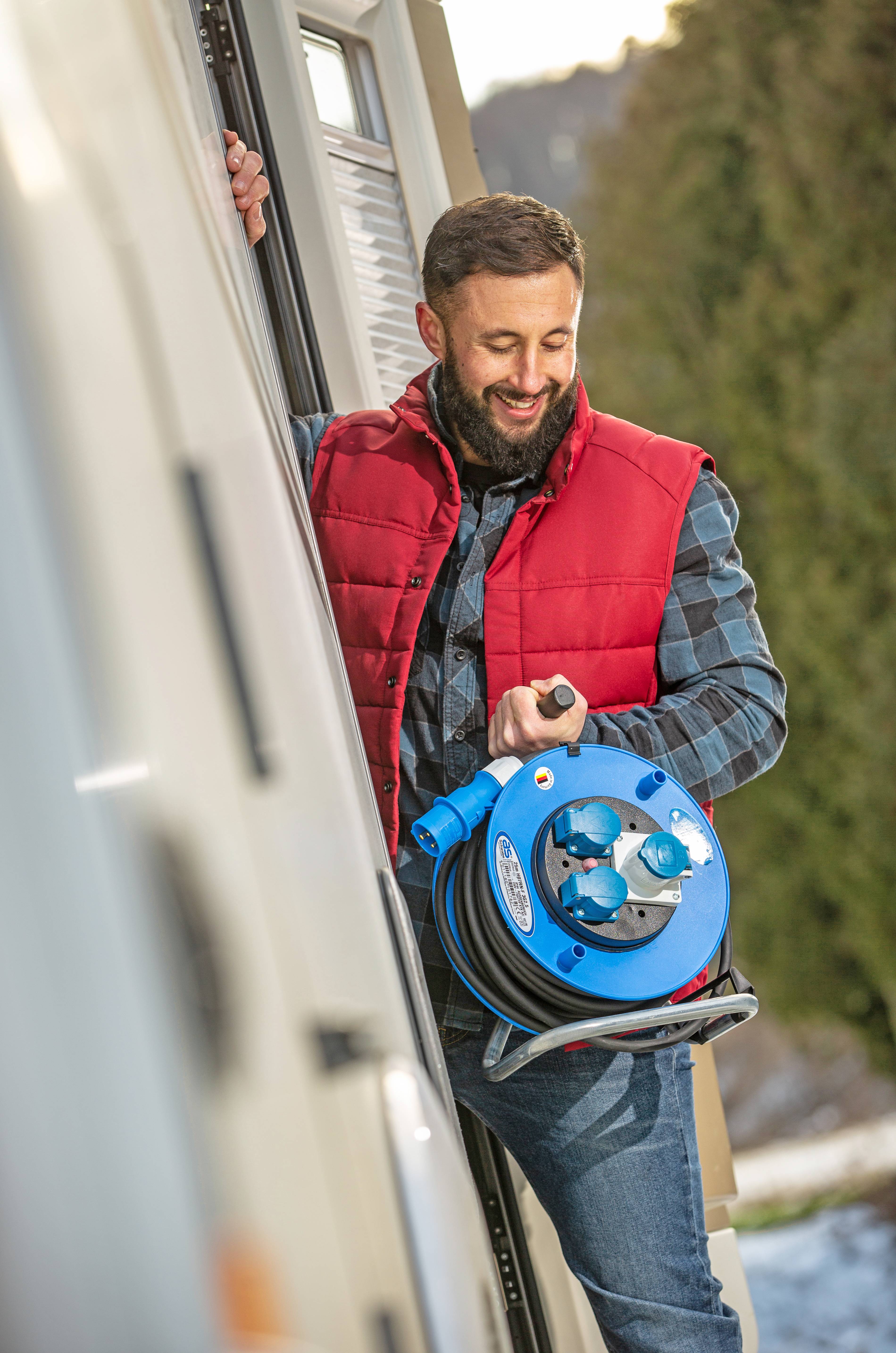 Een man in een rode bodywarmer draagt een blauwe kabelrol naast een camper. Op de achtergrond is vaag een haag te zien.