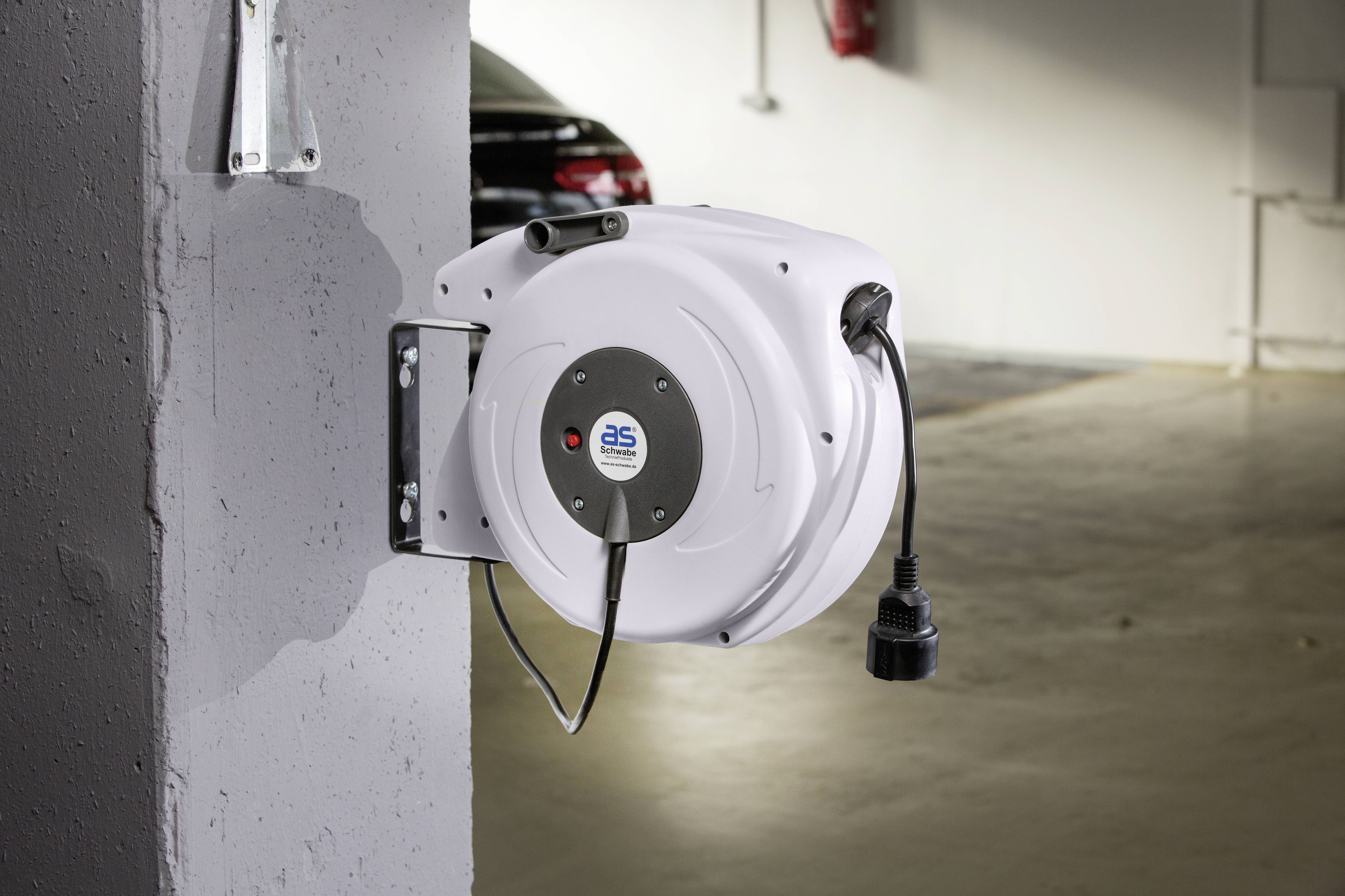Automatische kabeloproller gemonteerd aan een betonnen muur, met stroomkabel zichtbaar. Op de achtergrond gedeeltelijk zichtbare geparkeerde auto.