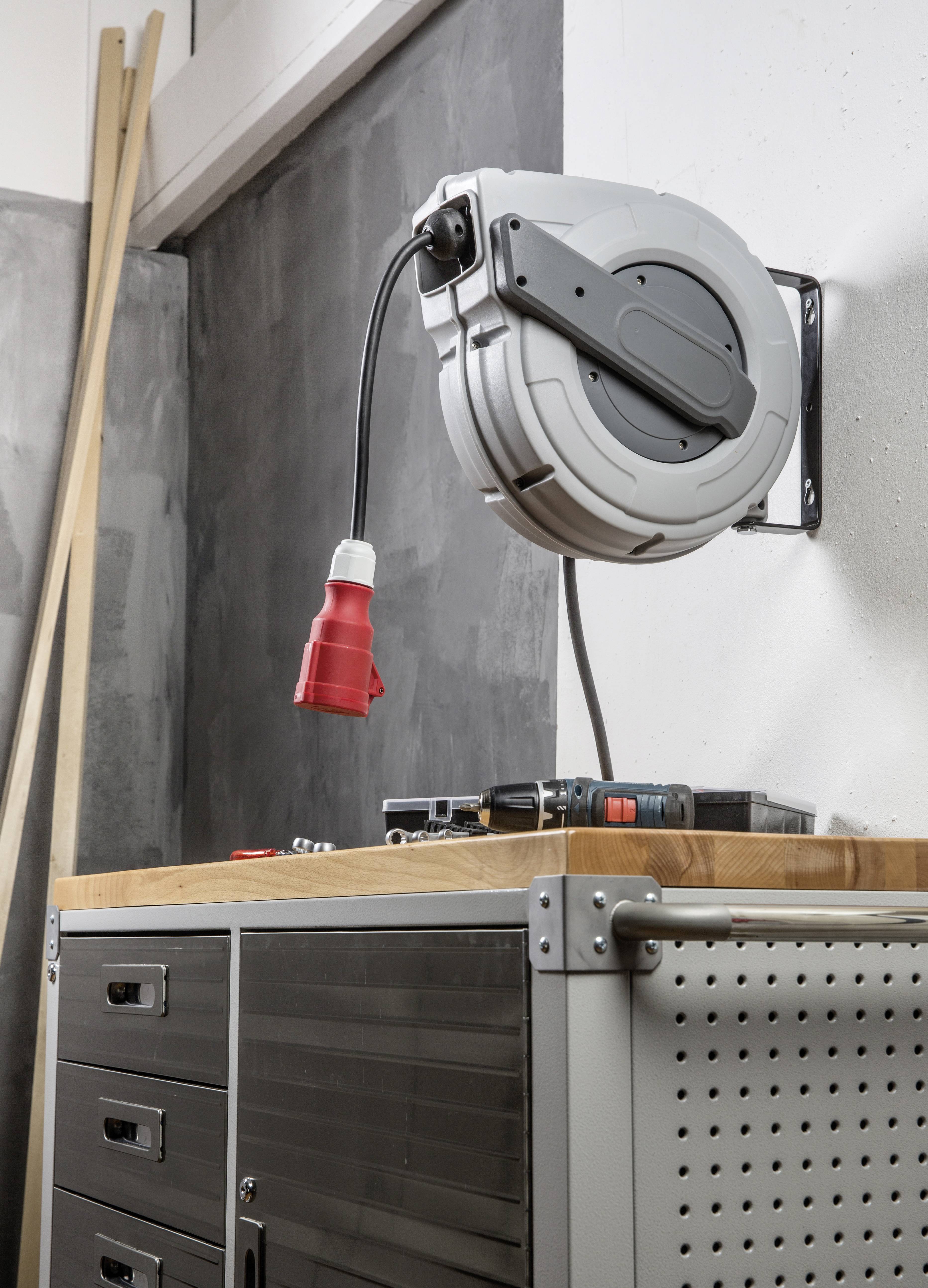 Een automatische kabeloproller is aan de muur gemonteerd boven een werkbank met laden en gereedschappen. Hij houdt een rood stroomsnoer vast.