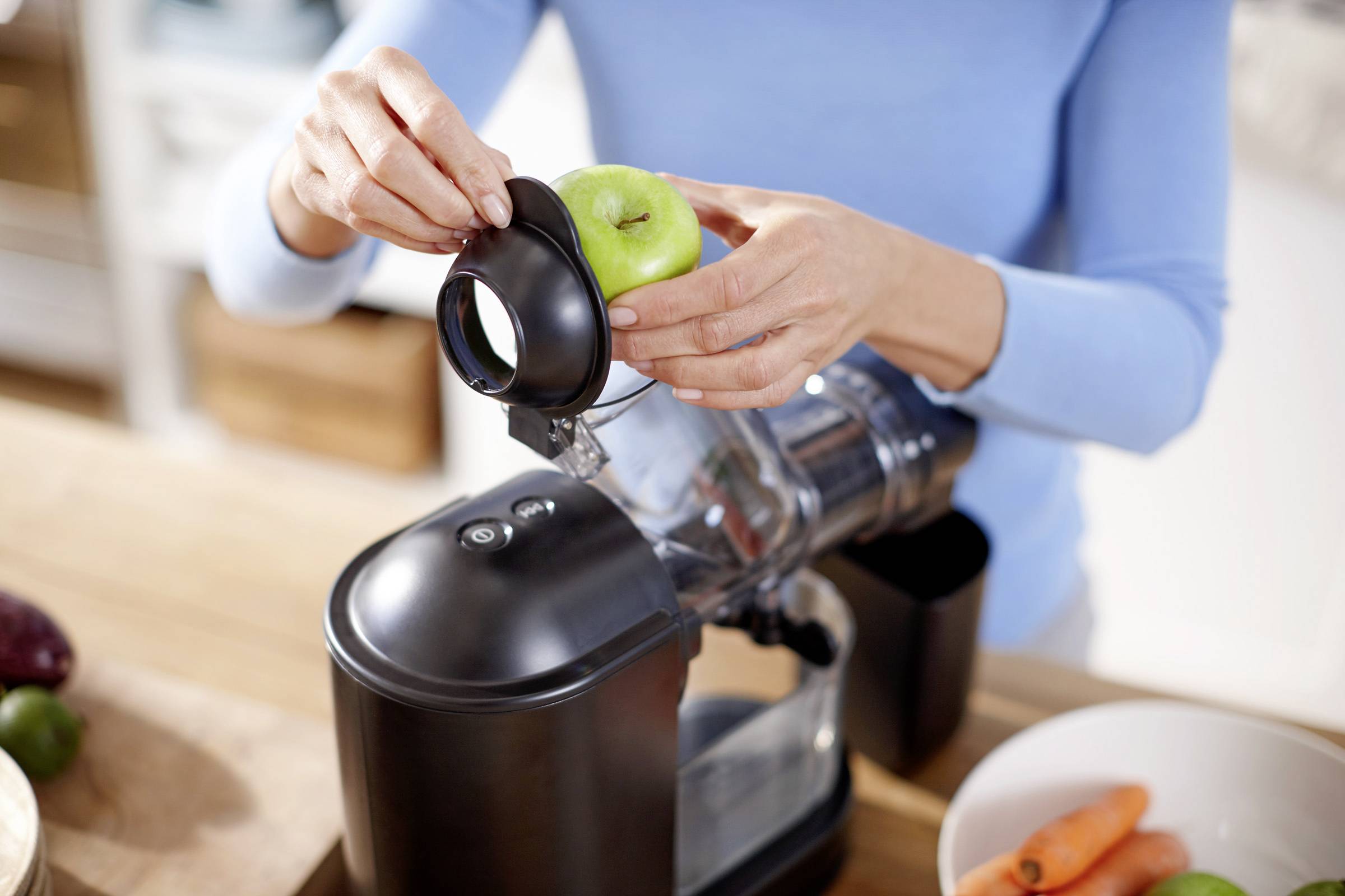 Een persoon stopt een groene appel in een zwarte sapcentrifuge in een keuken. Ernaast liggen wortels en ander fruit.