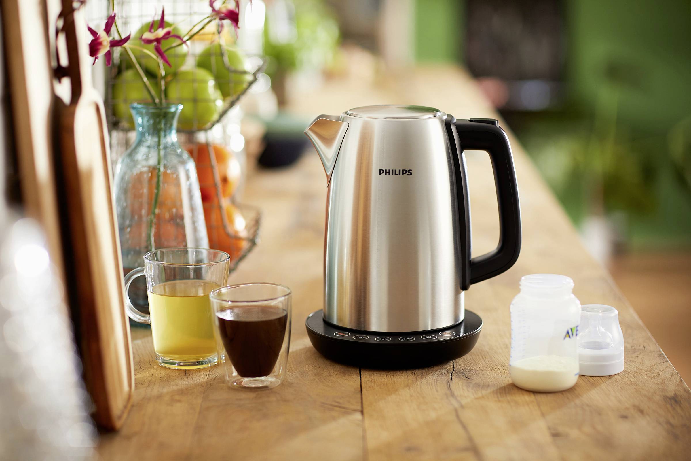 Een elektrische waterkoker op een houten tafel naast een glas sap, een kopje koffie en een flesje babymelk.