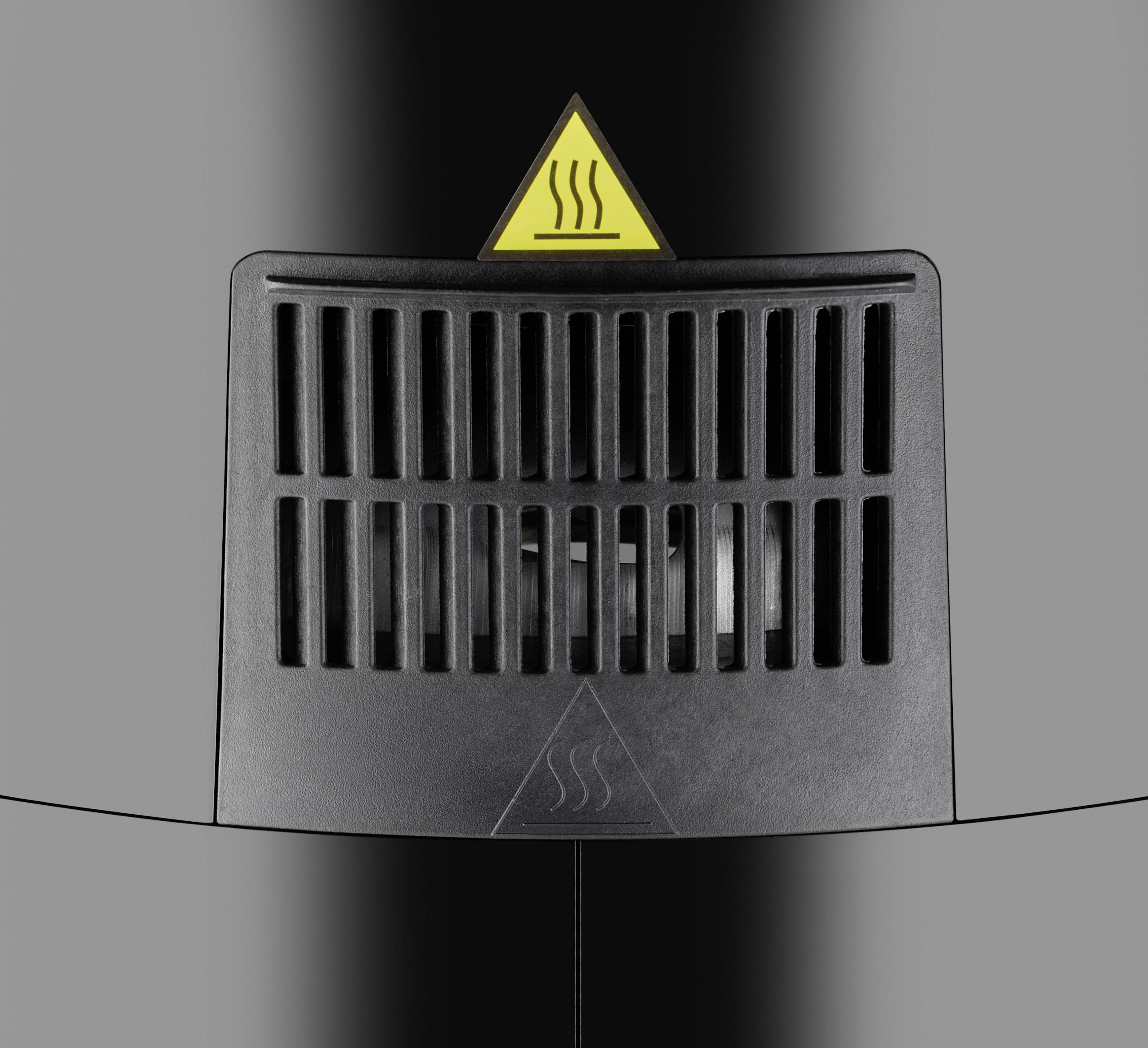 Zwart ventilatierooster op een grijze oppervlakte met een geel waarschuwingssymbool bovenaan dat hitte aangeeft.