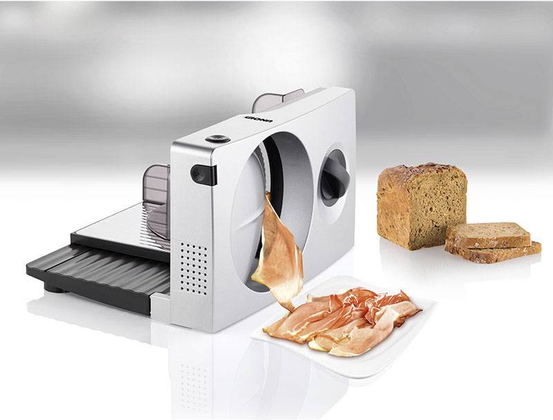 Elektrische snijmachine met een bord ham en twee plakken brood ernaast. Geschikt voor het snijden van brood, vlees en kaas.