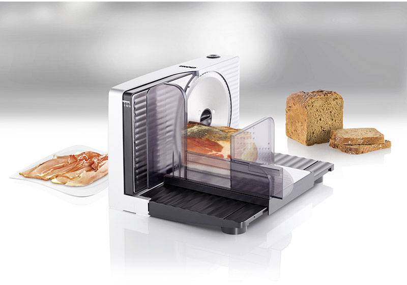 Elektrische snijmachine met brood en ham. Apparaat met zilveren behuizing, zwart snijvlak en transparante bescherming.