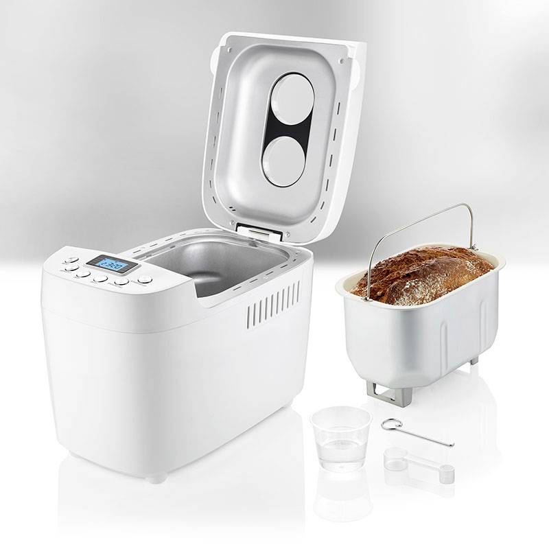 Broodbakmachine met open deksel, vers brood in de bakvorm ernaast. Toebehoren: maatbeker, haak, deeghaak.