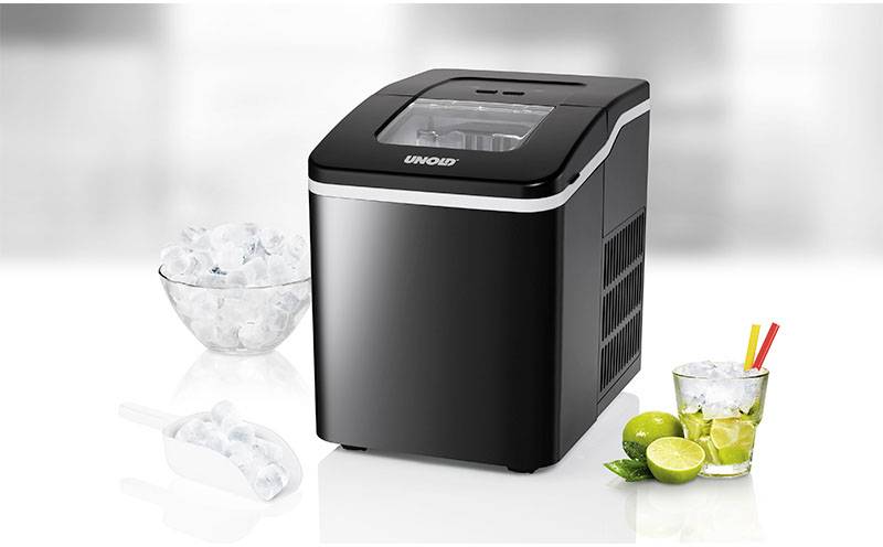 Zwarte ijsmachine op witte achtergrond, naast een kom met ijs, een ijsschep en een glas met limoenen en rietjes.