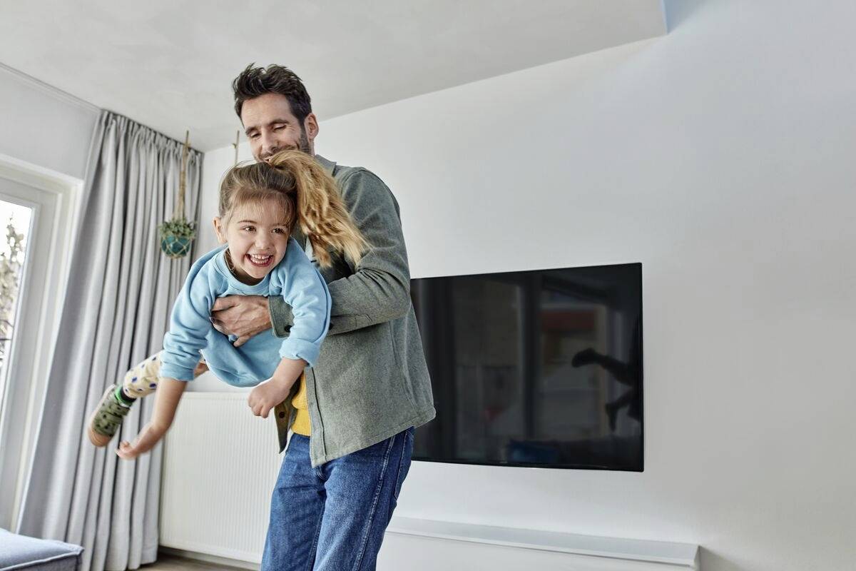 Een man tilt lachend een lachend kind op in de woonkamer. Op de achtergrond zijn een televisie en een raam met gordijnen zichtbaar.