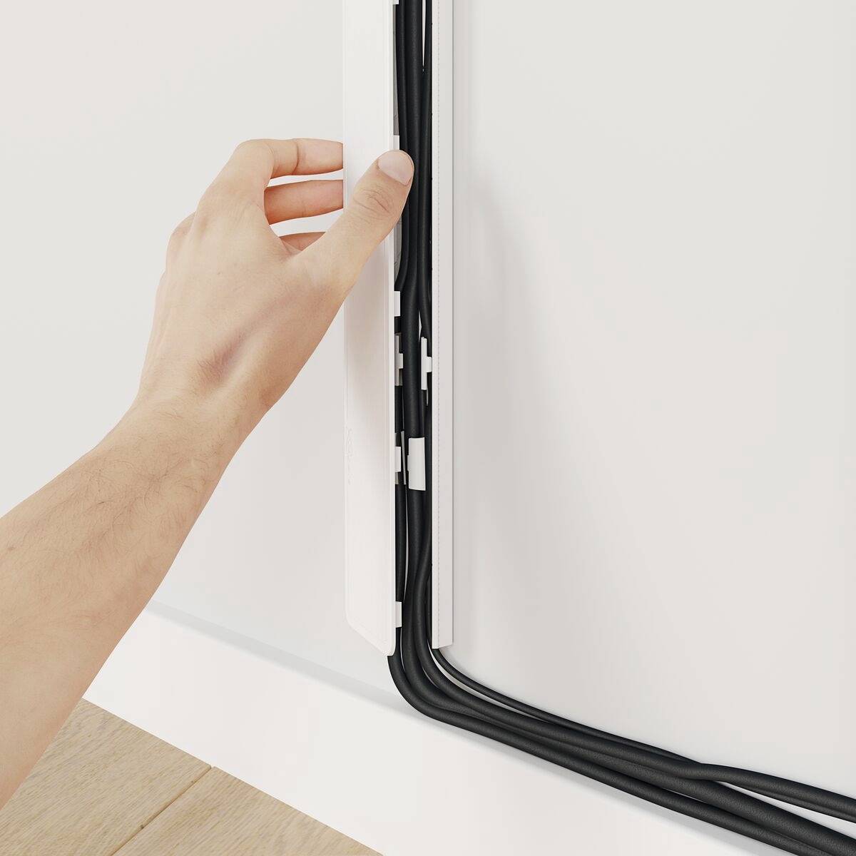 Een hand schuift verschillende zwarte kabels in een wit kabelkanaal langs een muur om de kabels netjes te verbergen.