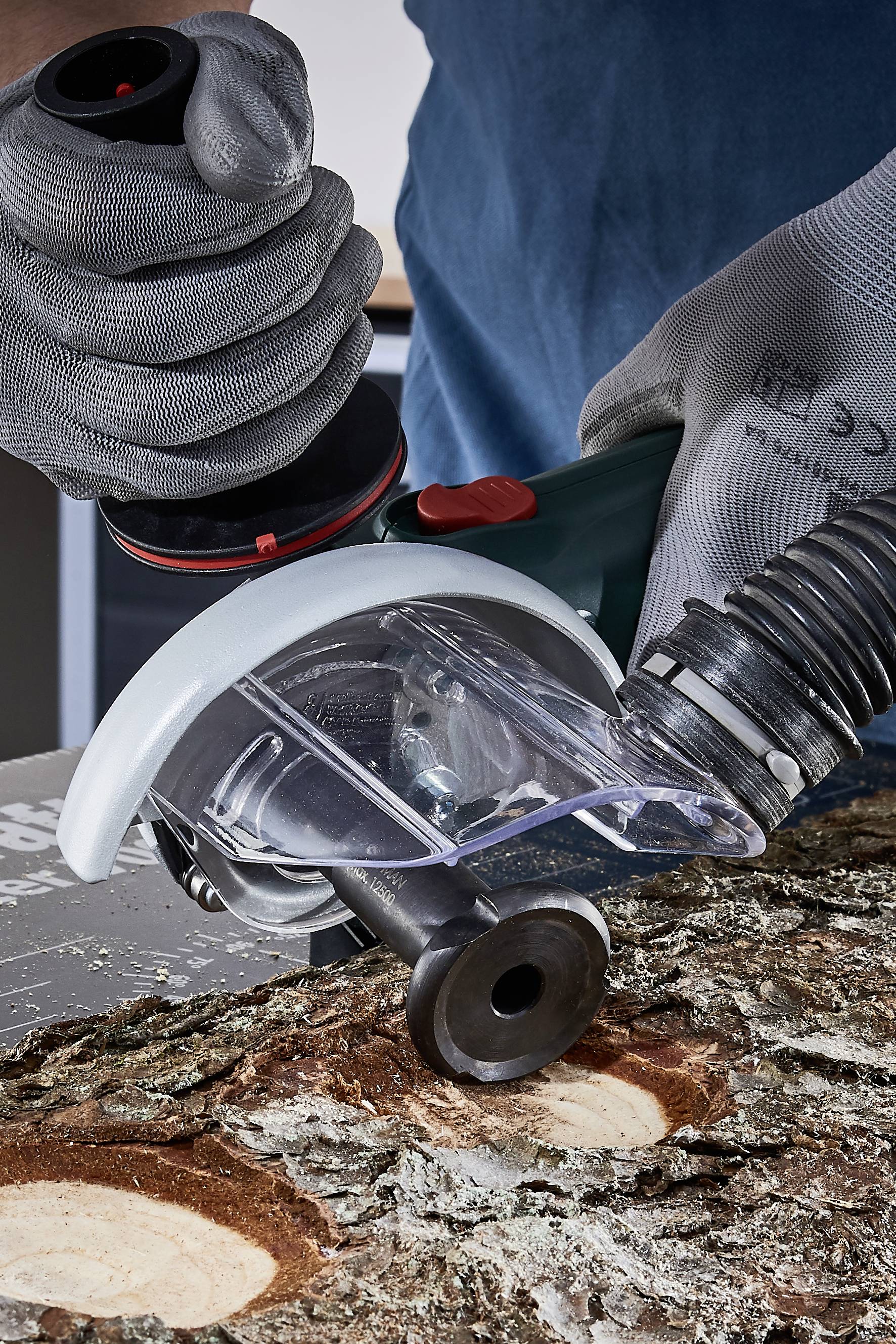 Een persoon draagt beschermende handschoenen en houdt een elektrisch gereedschap vast dat hout bewerkt. Houtspaanders en boomschors zijn zichtbaar.
