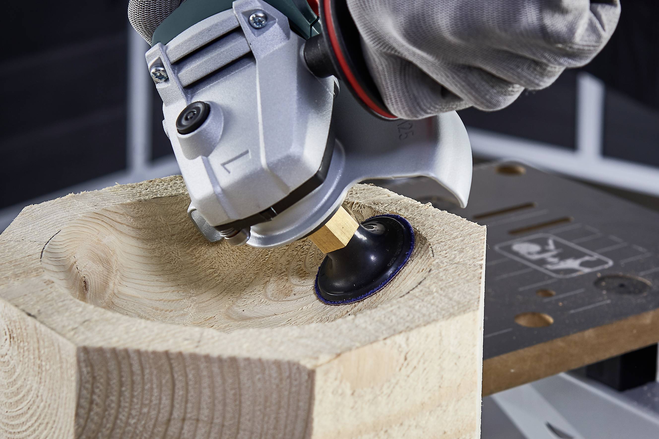 Een hand houdt een elektrisch gereedschap vast en bewerkt het oppervlak van een houten blok om het glad te maken. Planken zijn zichtbaar op de achtergrond.