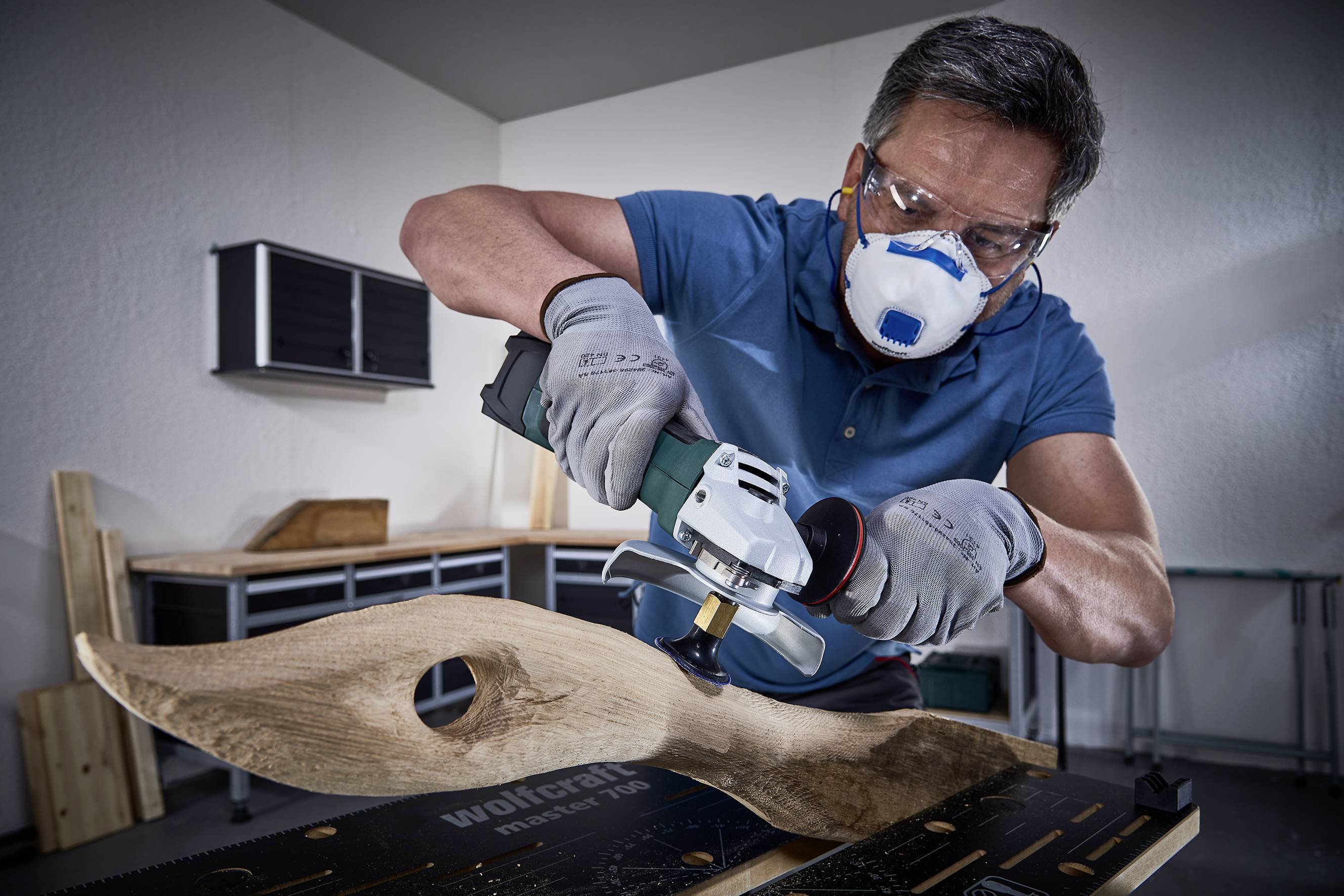 Een man slijpt een houten sculptuur met een elektrisch gereedschap in een werkplaats. Hij draagt een veiligheidsbril en een ademhalingsmasker.