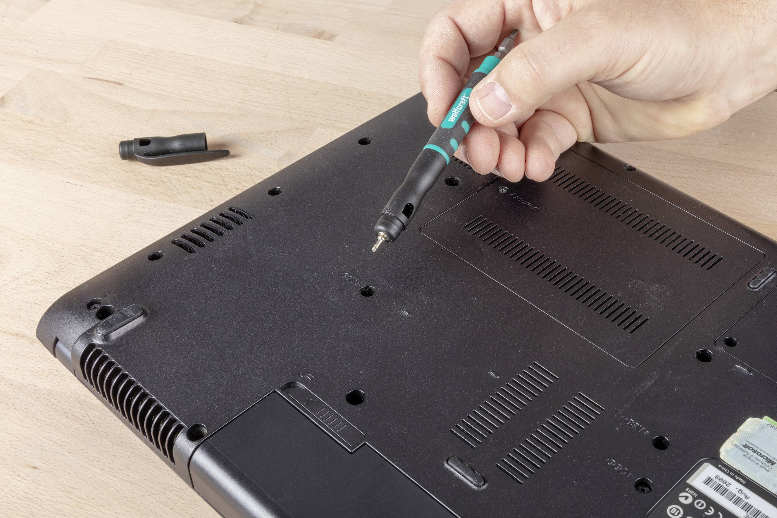 Een hand houdt een schroevendraaier vast en verwijdert een schroef van de achterkant van een elektronisch apparaat op een houten tafel.