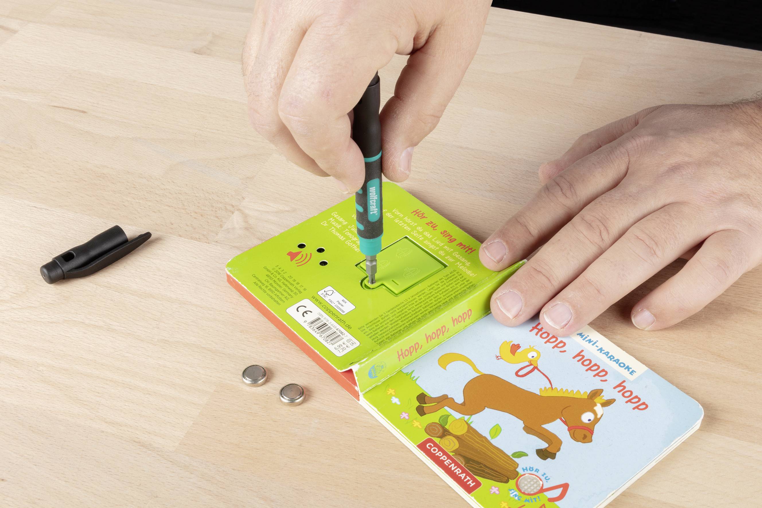 Een persoon werkt met een klein boek en een schroevendraaier op een tafel, waarbij losse batterijen en een penndop ernaast liggen.