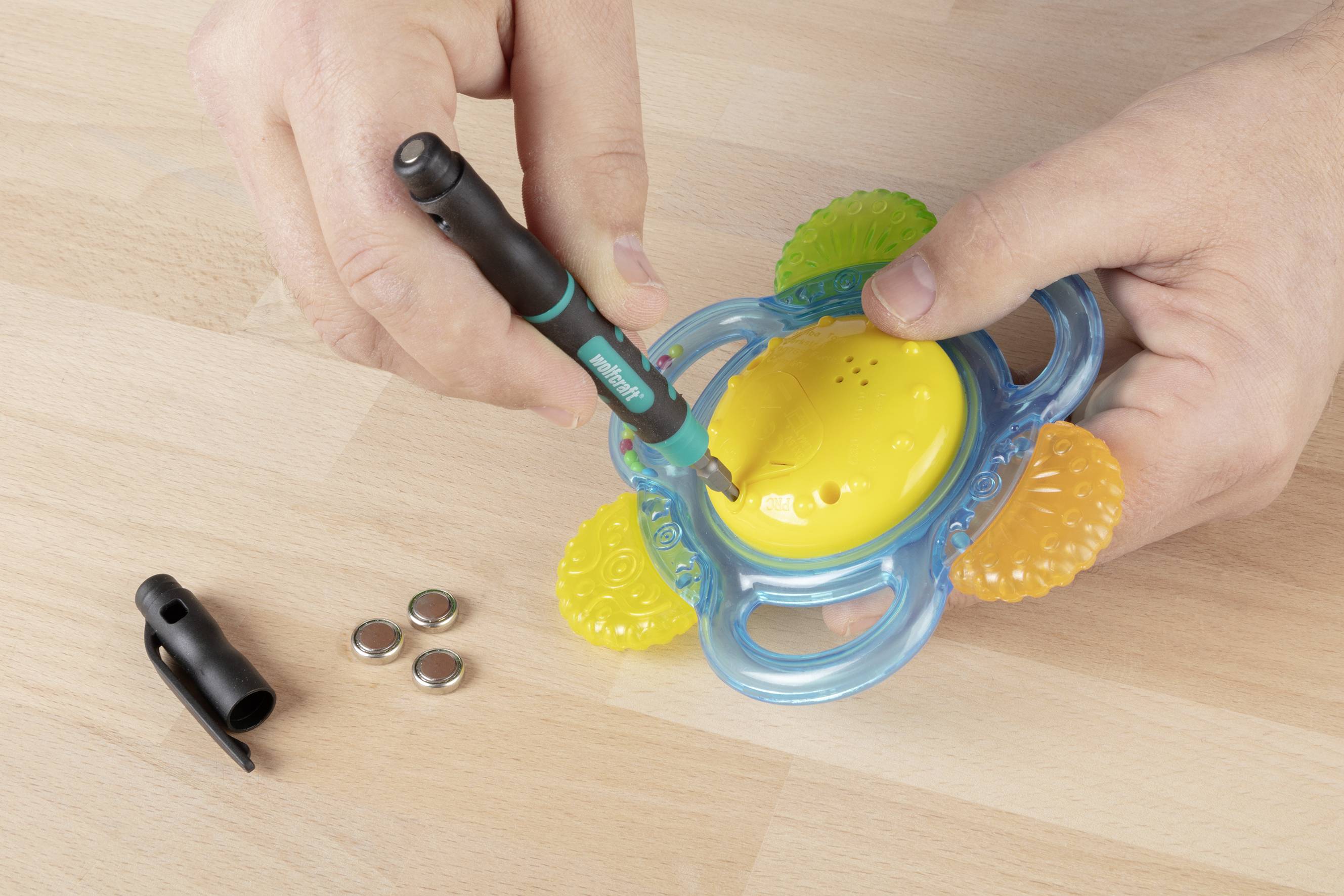 Een persoon demonteert een geel, rond speelgoed met blauwe handgrepen en kleurrijke details met een kleine schroevendraaier.