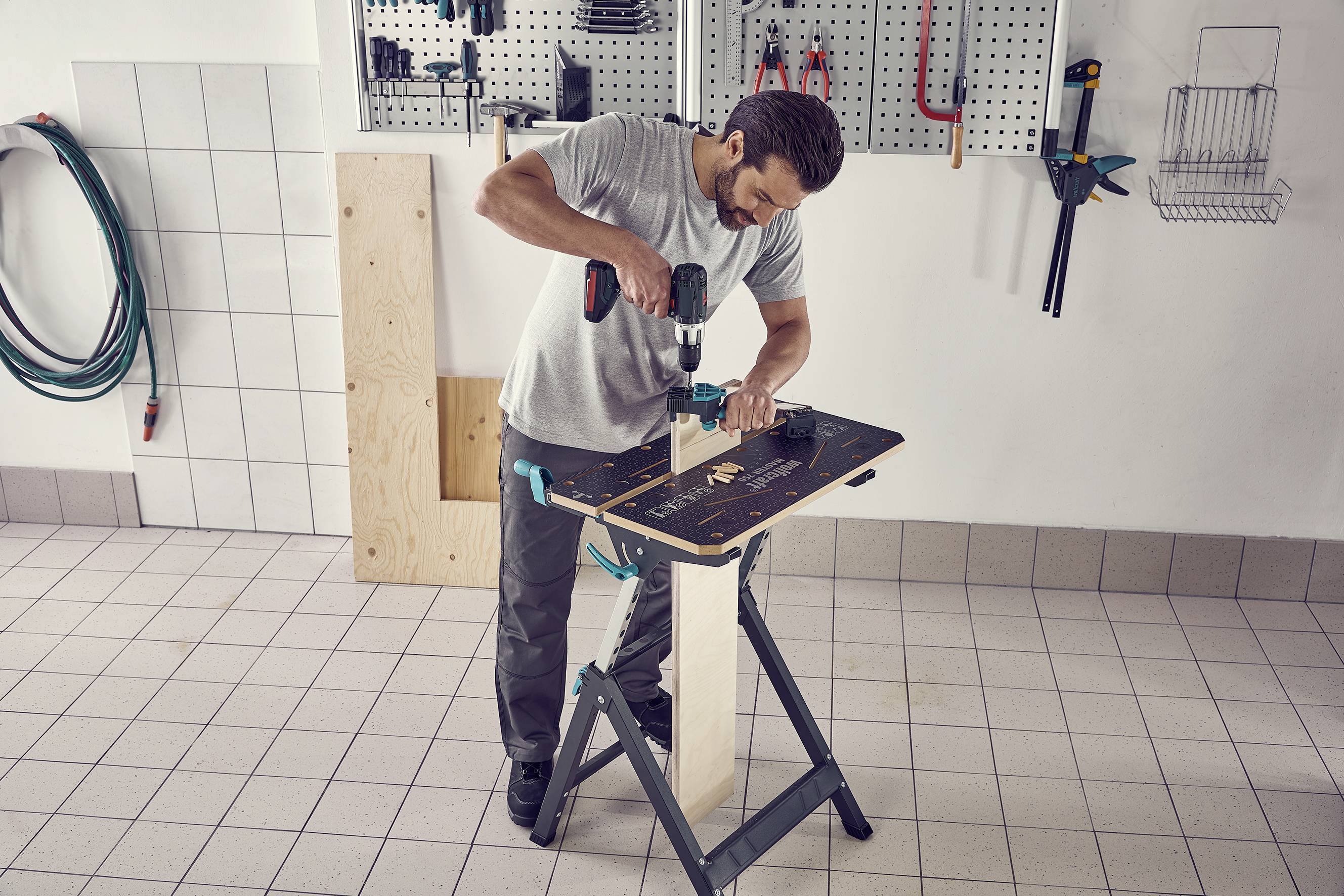 Een man werkt in een werkplaats. Hij boort met een accuschroevendraaier in een houten plaat die is vastgemaakt op een werkbank.
