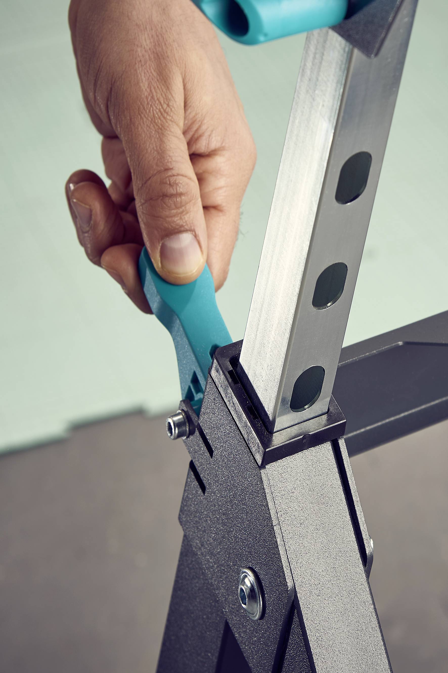 Een hand trekt aan een blauwe hefboom om de hoogte van een metalen frame met meerdere gaten aan te passen.