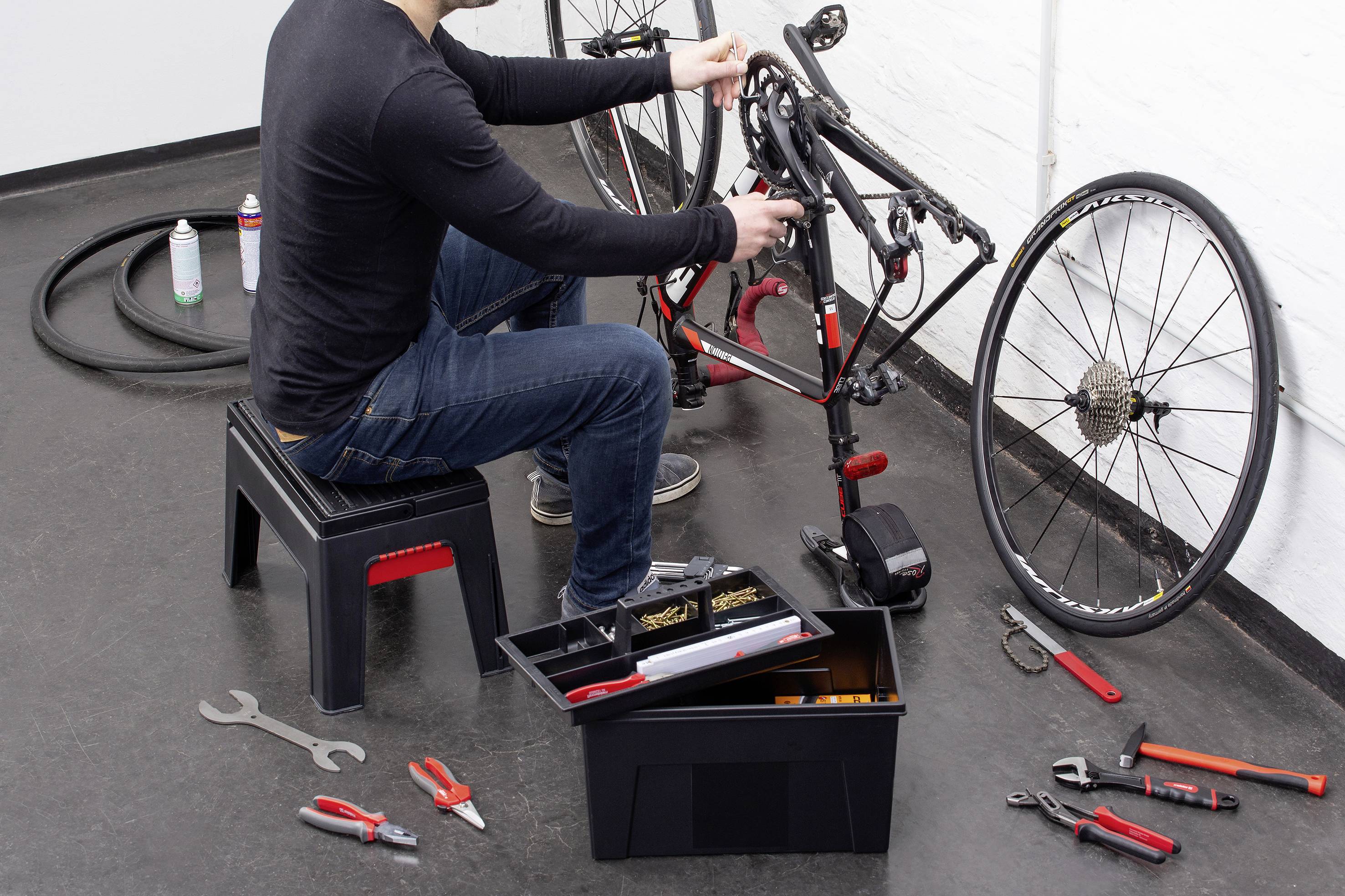 Een persoon repareert een fiets die op een standaard staat. Gereedschappen en een gereedschapskist liggen om haar heen op de grond.