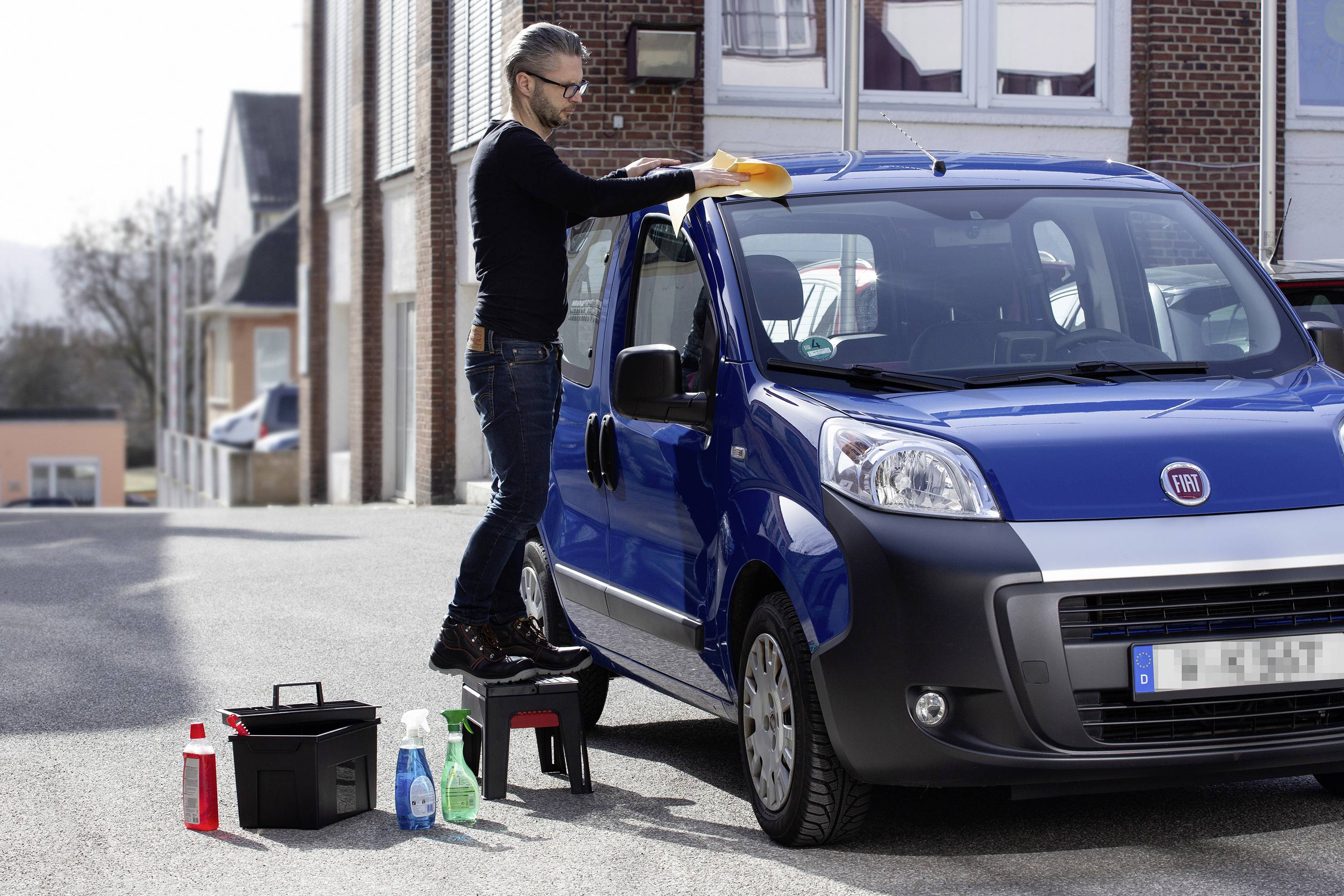 Een man staat op een kleine trap en poetst het dak van een blauwe bestelwagen. Verschillende schoonmaakmiddelen staan ernaast op de grond.