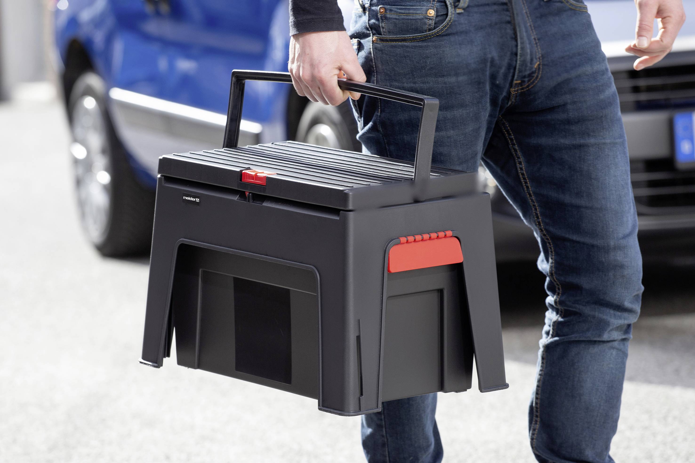 Een persoon draagt een zwarte, stevige transportbox met een rode sluiting. Op de achtergrond is een onscherpe blauwe auto zichtbaar.