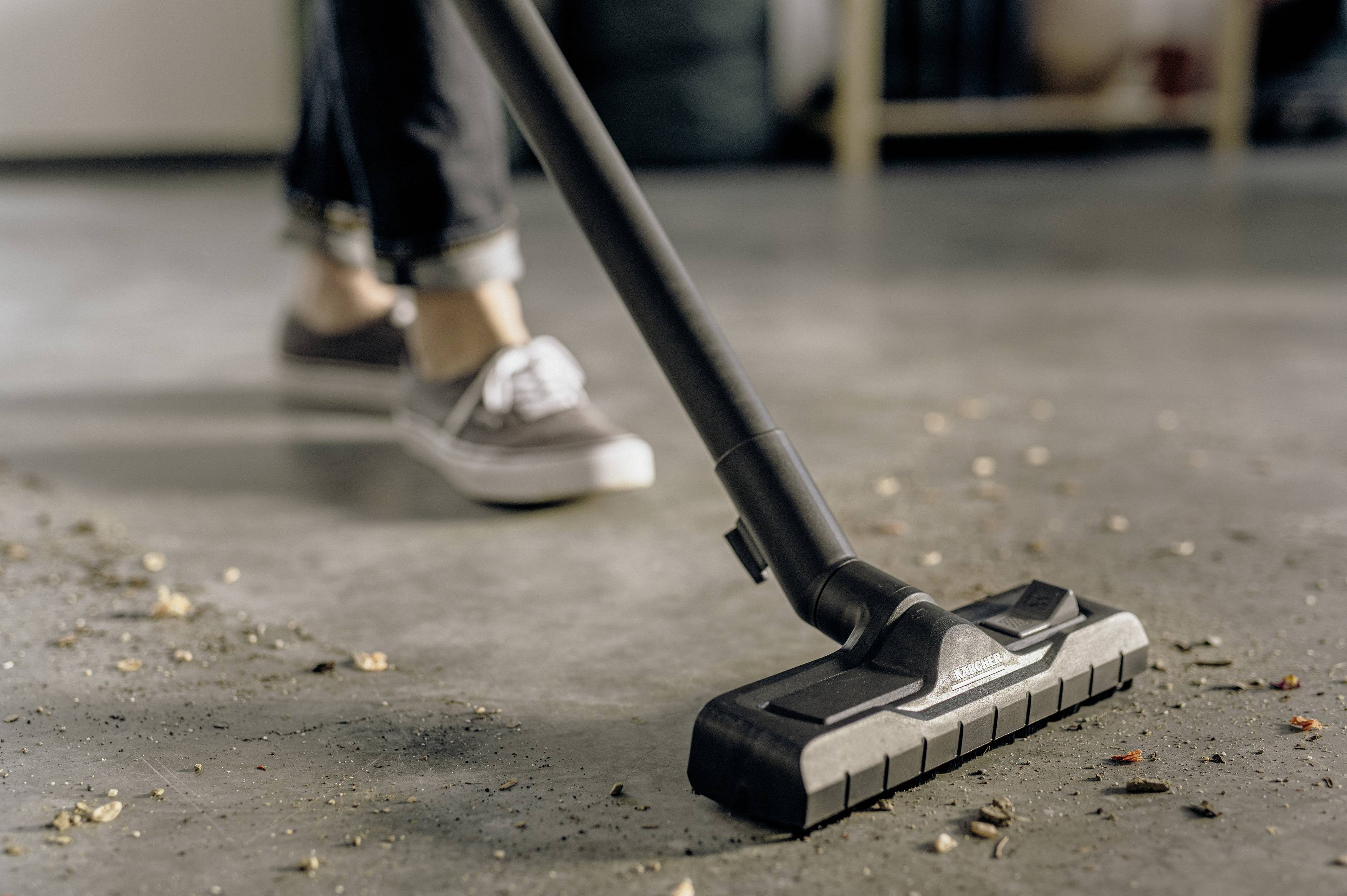 Een persoon zuigt met een stofzuiger een vuile vloer. Men ziet alleen de schoenen en de stofzuiger, terwijl vuil wordt verwijderd.