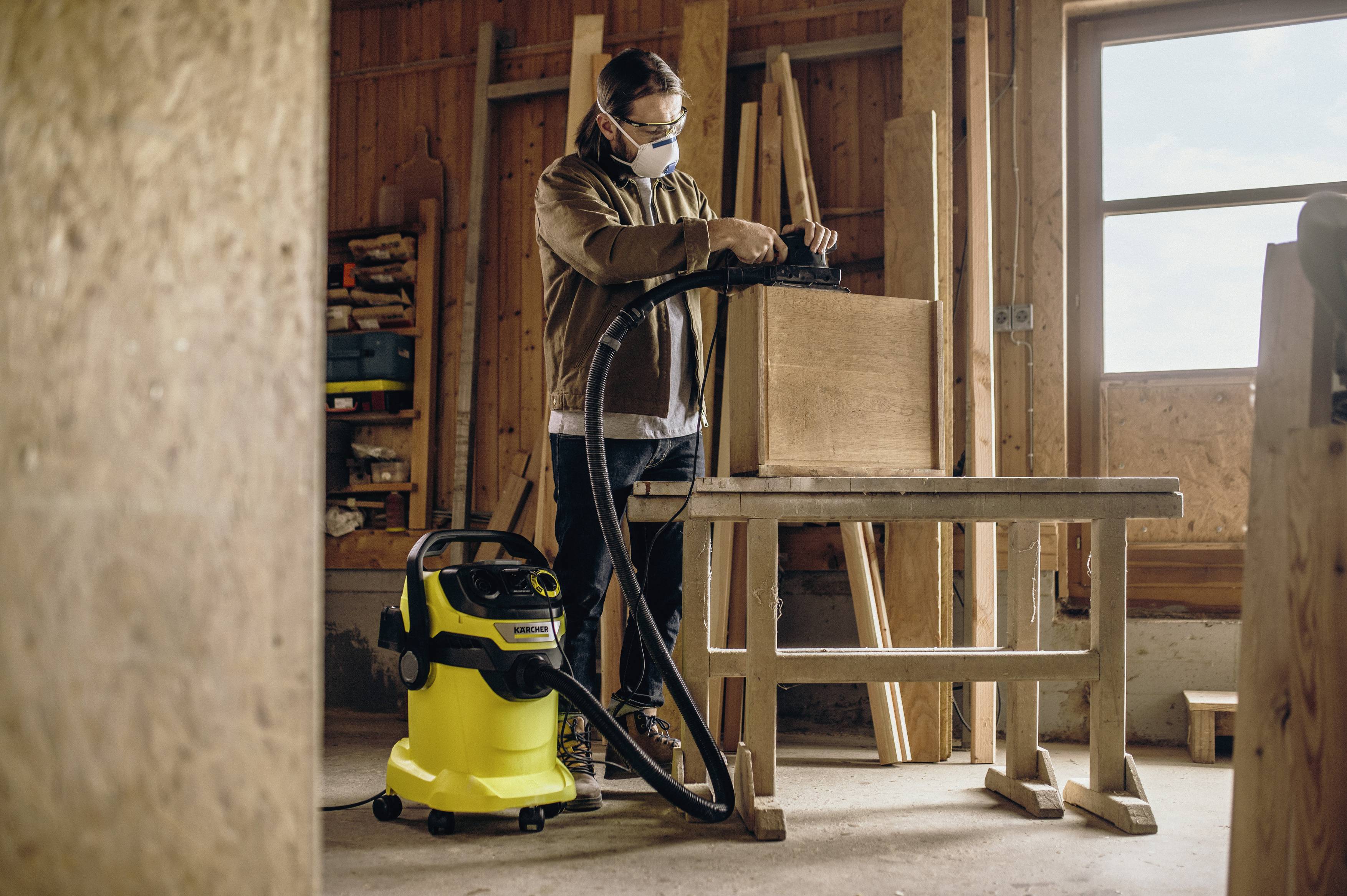 Een timmerman werkt in een werkplaats en gebruikt een stofzuiger om houtspaanders van een werktafel te verwijderen.