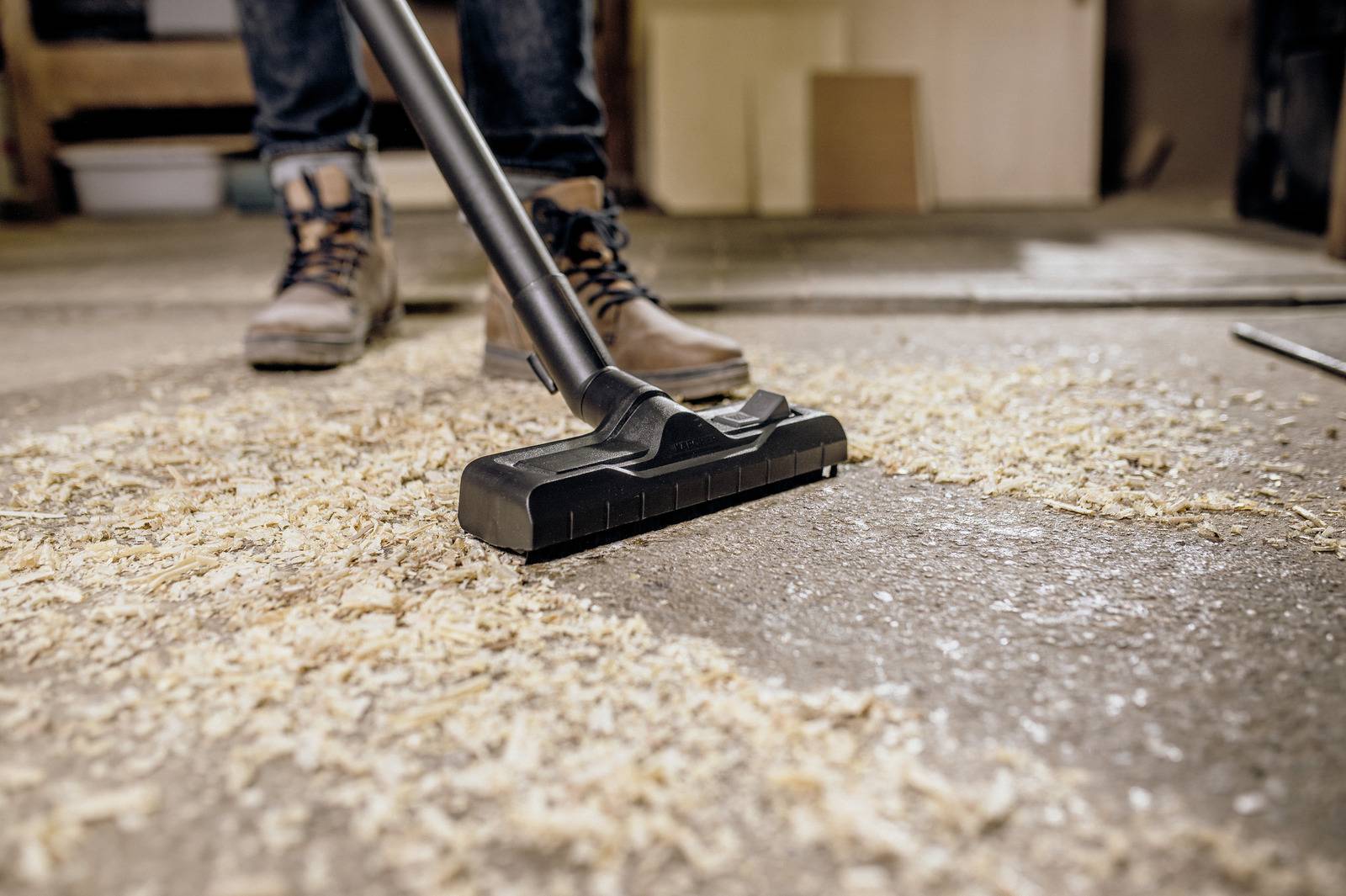 Een persoon draagt werkschoenen en gebruikt een stofzuiger om zaagsel van een betonnen vloer te reinigen, wat wijst op een werkplaats of bouwkundige omgeving.