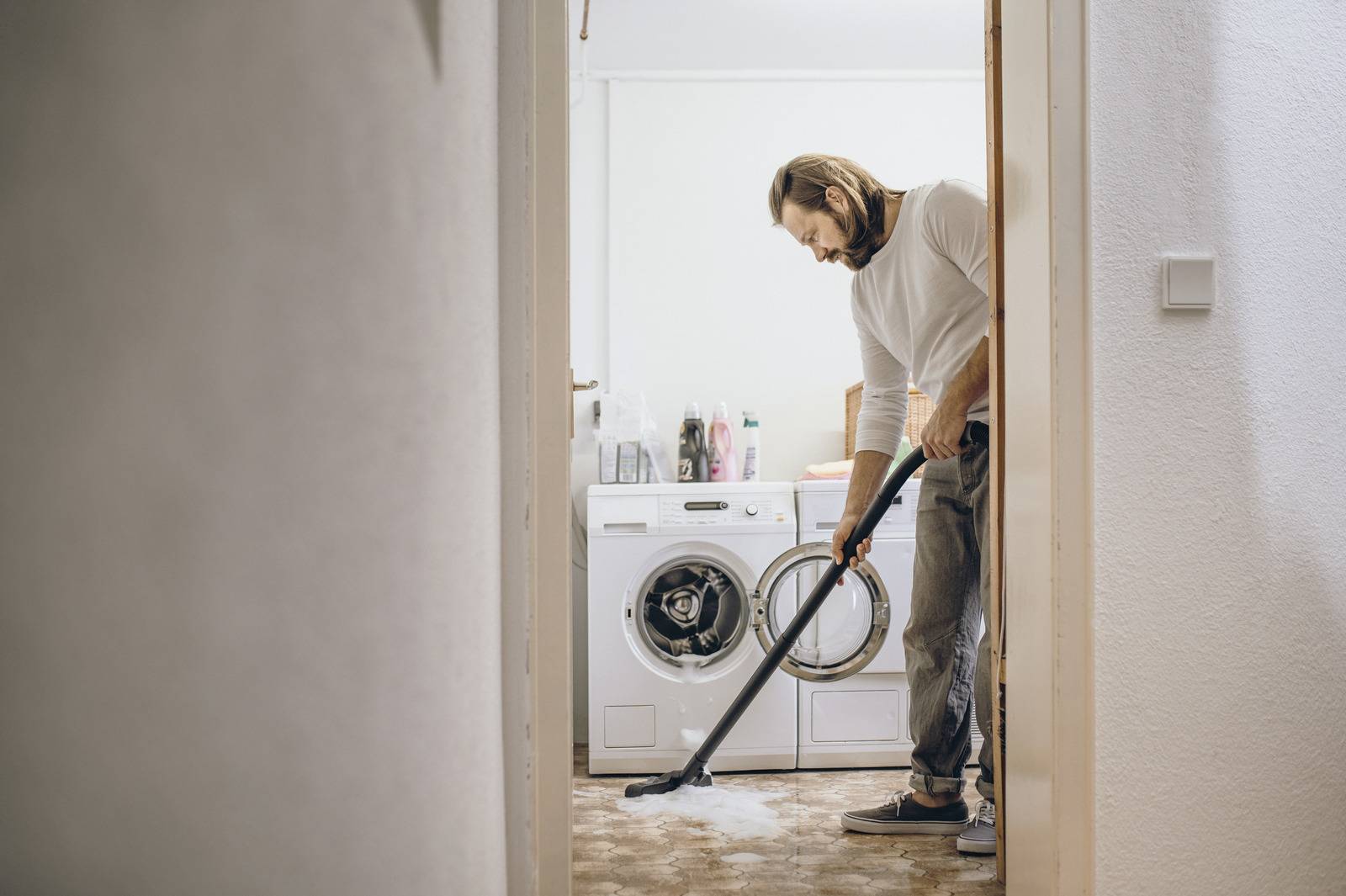 Een persoon die de vloer van een wasruimte stofzuigt, naast een wasmachine en droger.