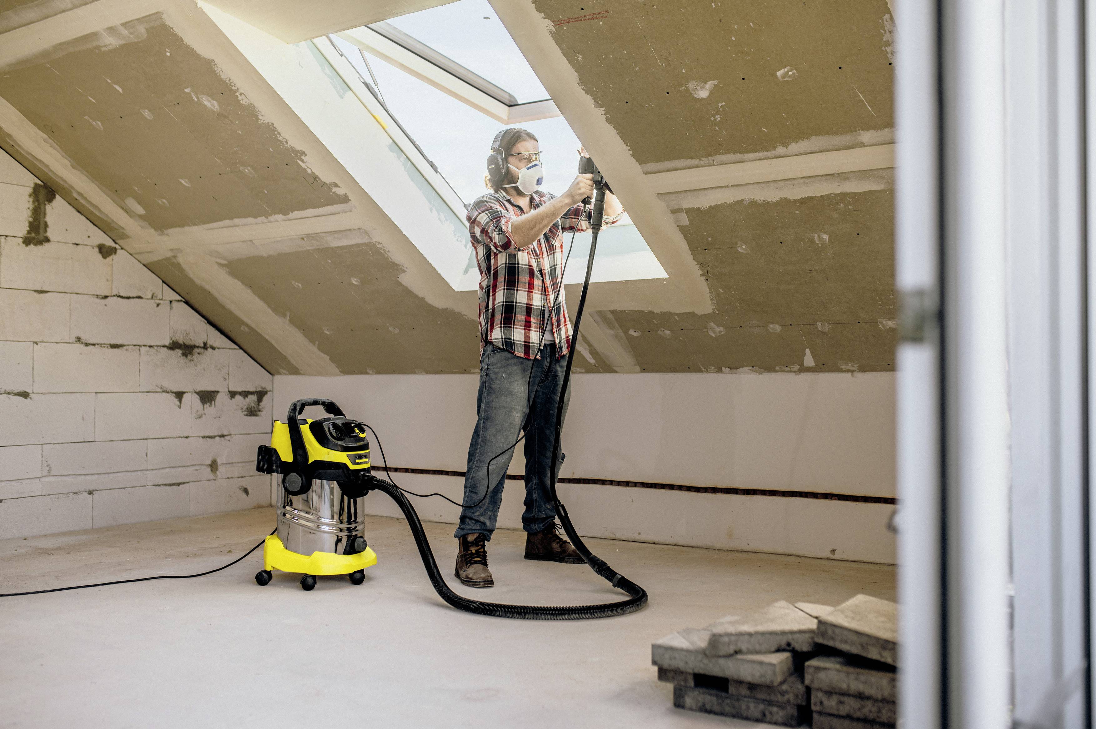 Een persoon reinigt een zolderkamer met een industriële stofzuiger, terwijl een groot dakraam het vertrek van natuurlijk licht voorziet.