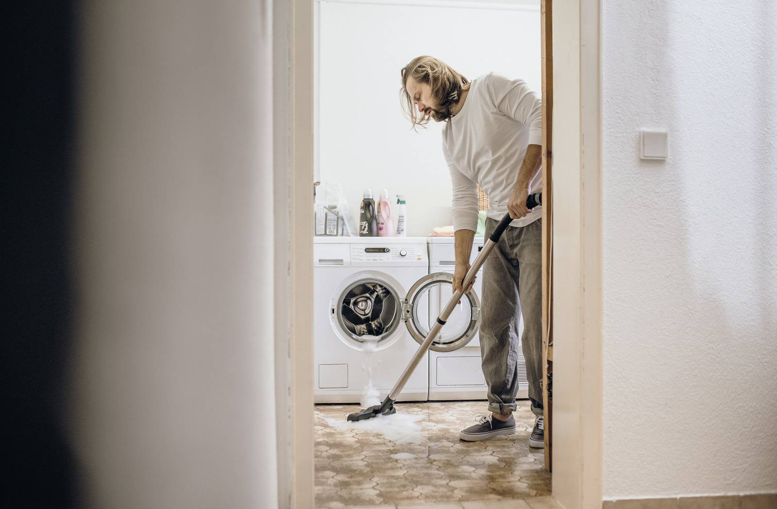 Een persoon zuigt een tapijt met een stofzuiger in een wasruimte met een wasmachine, droger en schoonmaakmiddelen zichtbaar op de achtergrond.