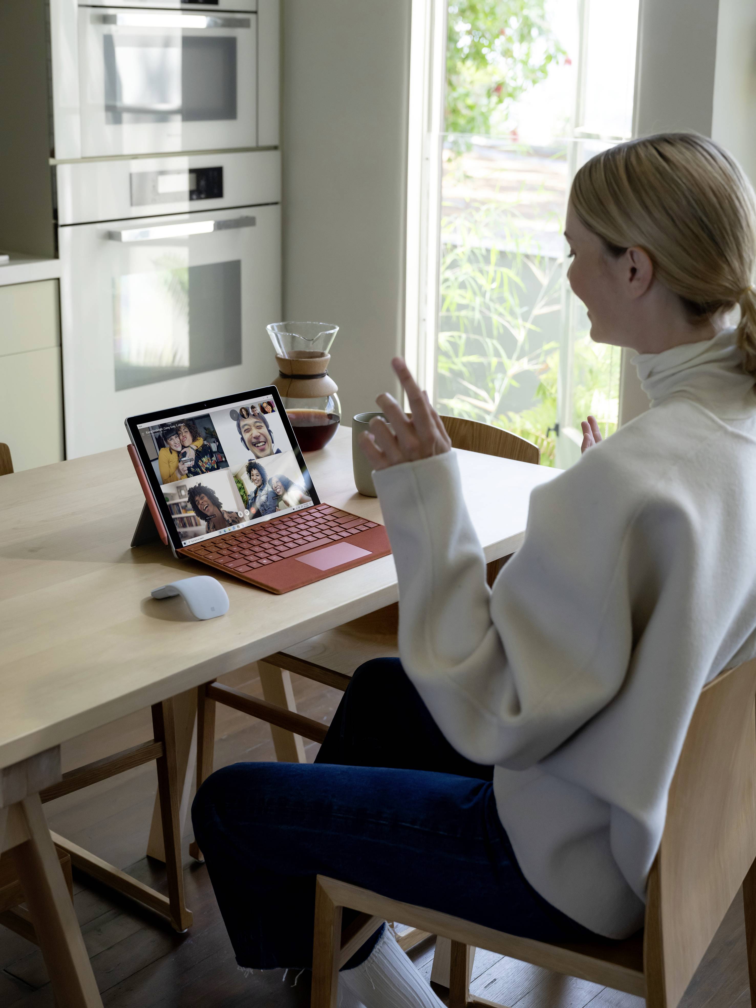 Een persoon zit aan tafel en neemt deel aan een videoconferentie op een laptop. Op de achtergrond zijn een kachel en planten zichtbaar.