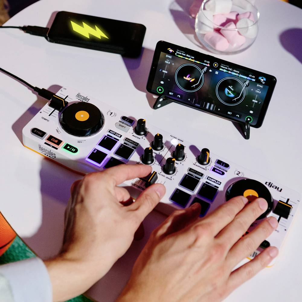 Een persoon bedient een DJ-mengpaneel met draaiknoppen. Een smartphone vertoont een muziek-mix-app. De setup staat op een tafel.