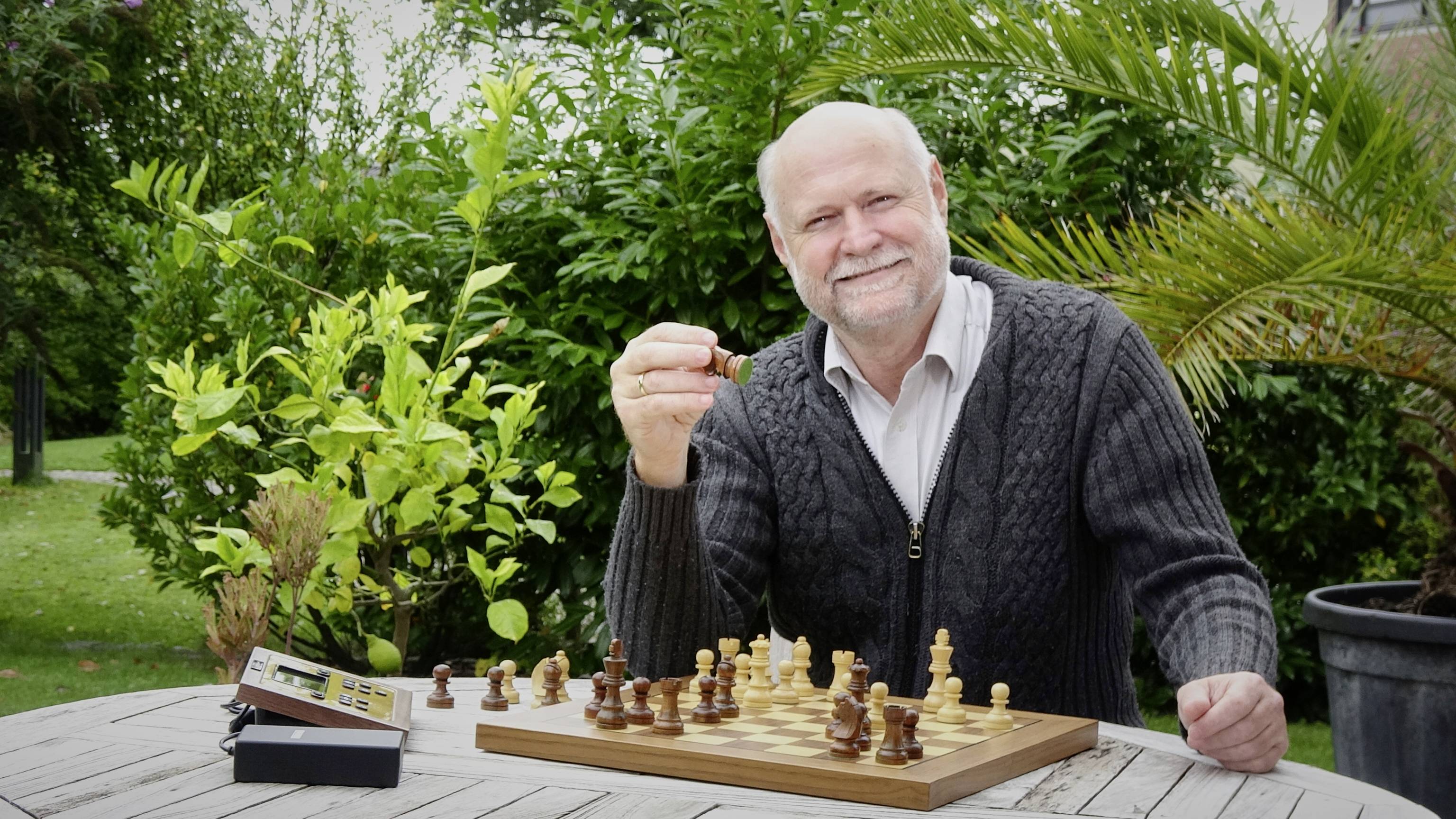 Een oudere man zit glimlachend in de tuin aan een tafel met een schaakbord. Hij houdt een schaakstuk in zijn hand.