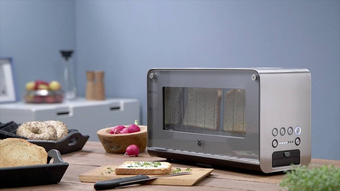 Een zilveren broodrooster op een houten tafel, naast een snijplank met brood, een mes en een kom met radijsjes.