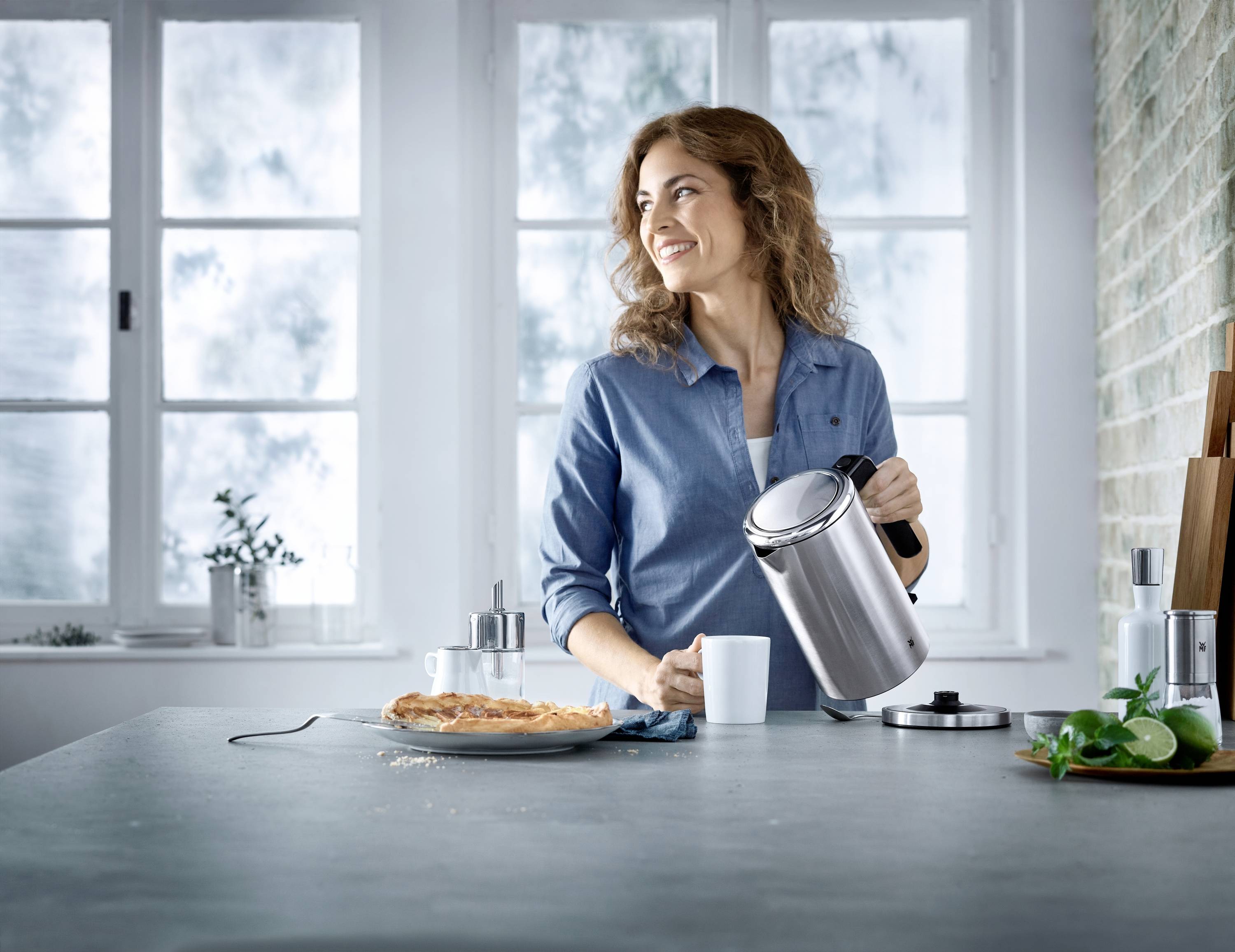 Een vrouw in een blauw shirt giet water uit een waterkoker in een kopje. Op tafel liggen een cake en keukengerei.