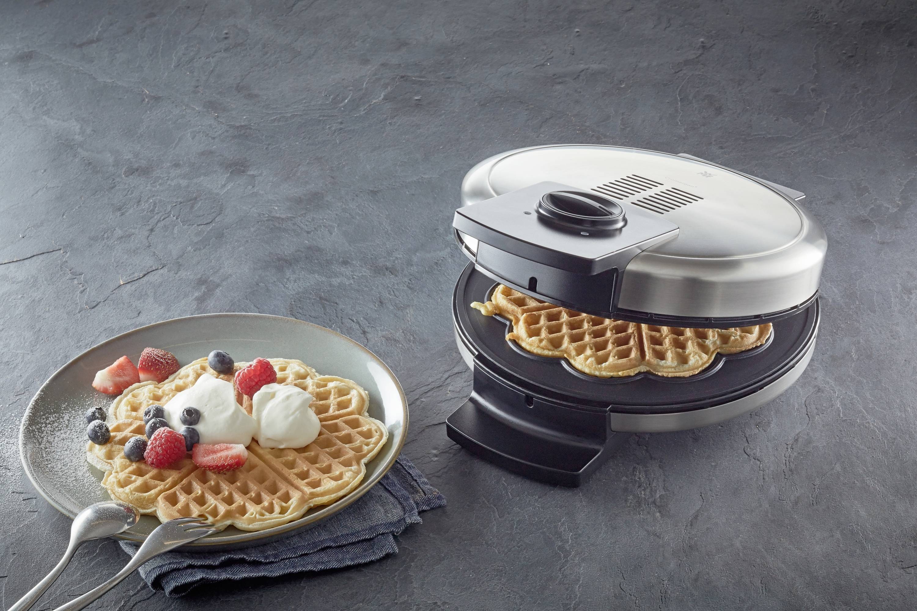 Een wafelijzer met wafels erin, naast een bord met een afgewerkte wafel, versierd met slagroom en bessen zoals aardbeien en bosbessen.