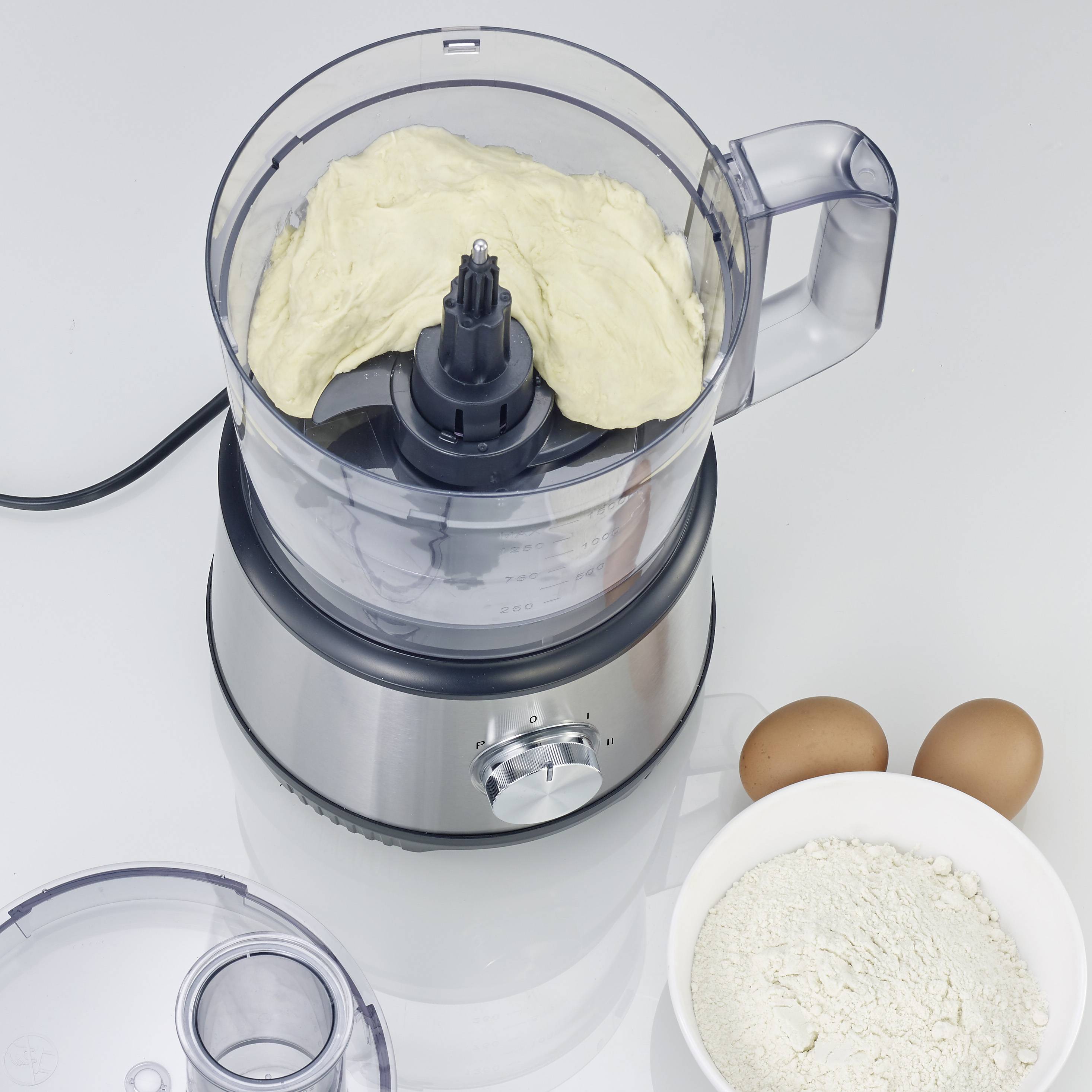 Een mixer met deegmassa erin, ernaast bloem in een kom en twee eieren op een tafel.