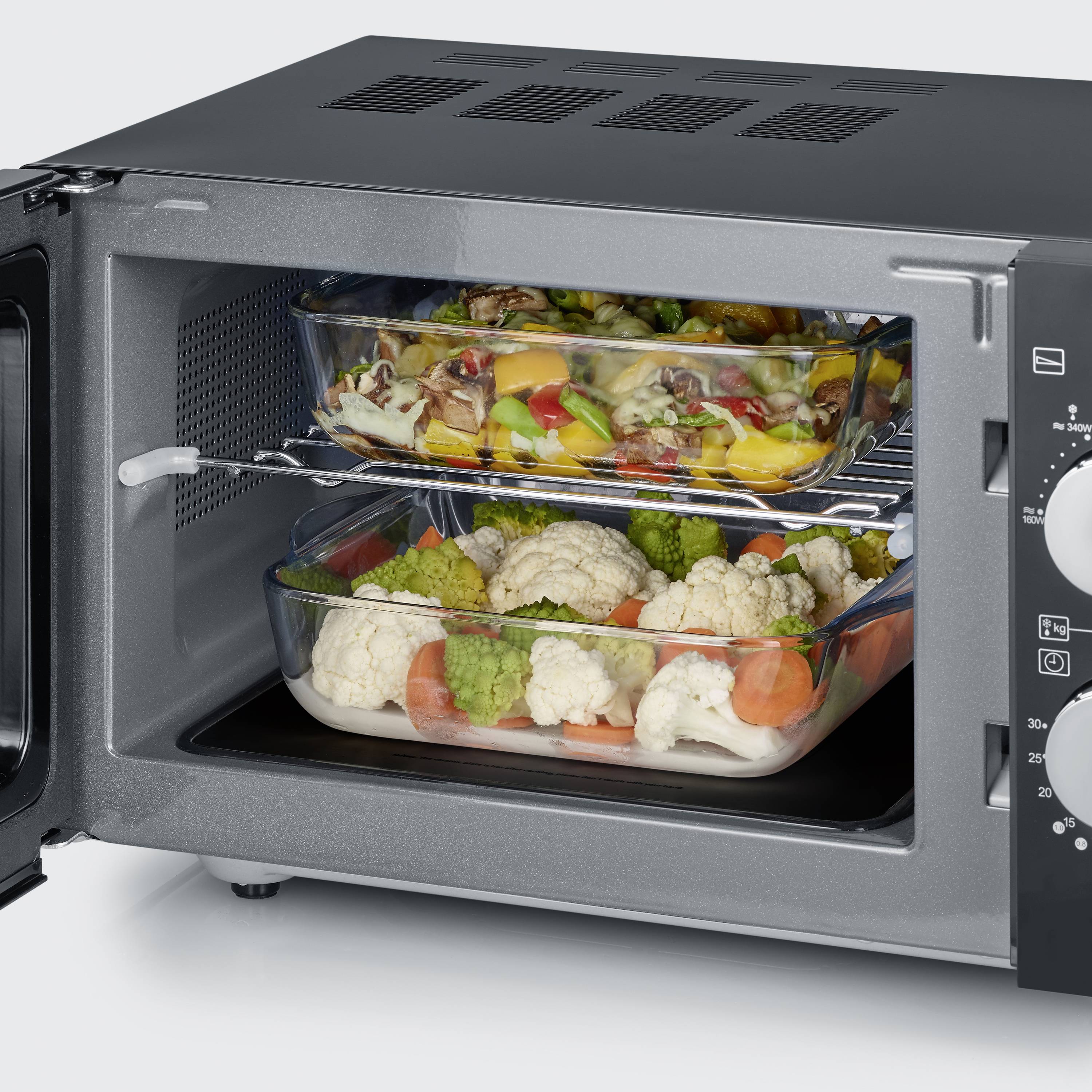 Een geopende magnetron toont twee glazen bakjes met groenten op de roosters. Daarin zijn kleurrijke stukken broccoli, bloemkool en paprika zichtbaar.