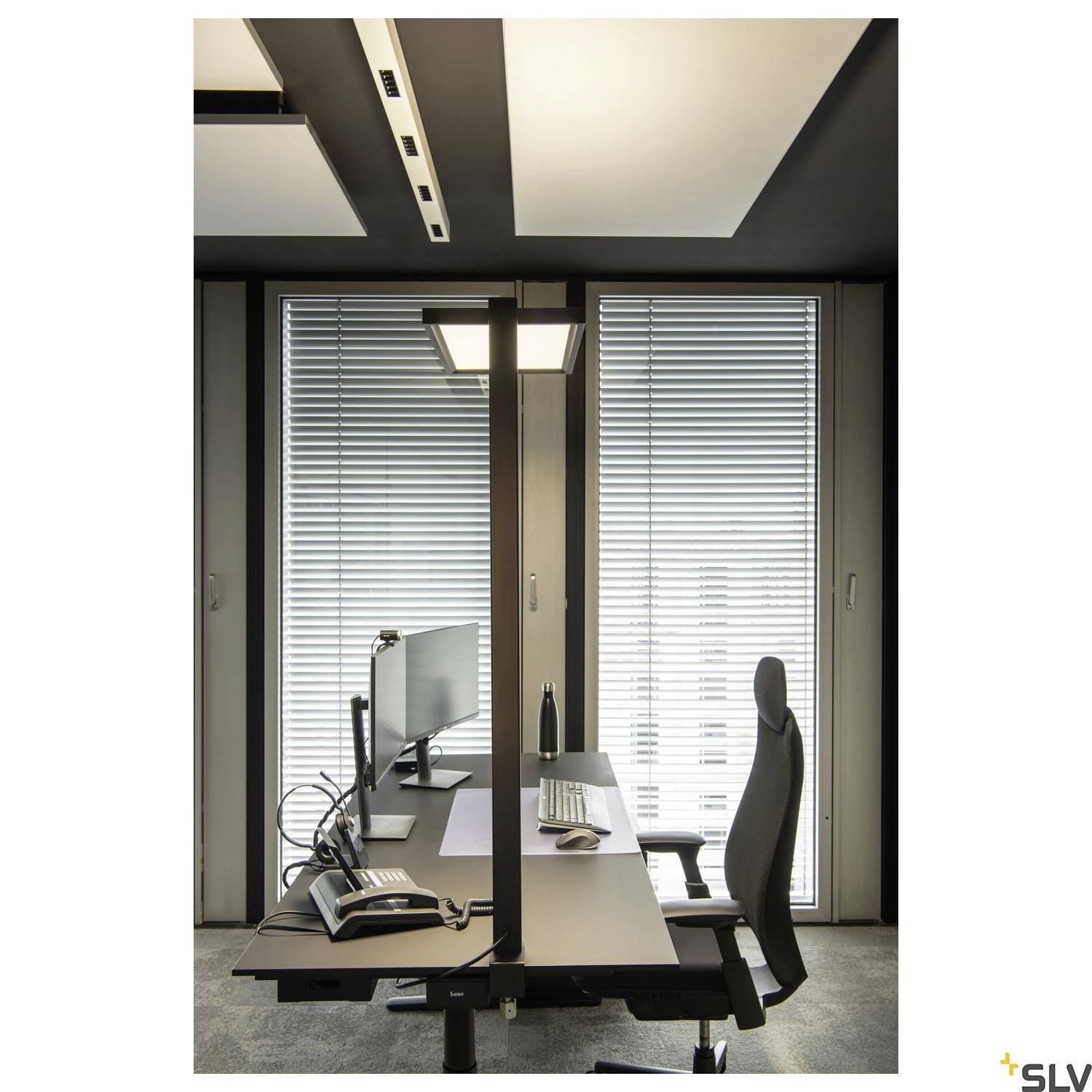 Een modern kantoor met twee bureaus, een computerbeeldscherm, een telefoon en ergonomische stoelen. Grote ramen met jaloezieën op de achtergrond.