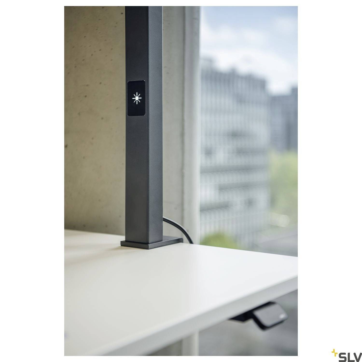 Een zwarte staande lamp met een vierkante voet staat op een witte tafel voor een raam. Op de achtergrond zijn gebouwen zichtbaar.