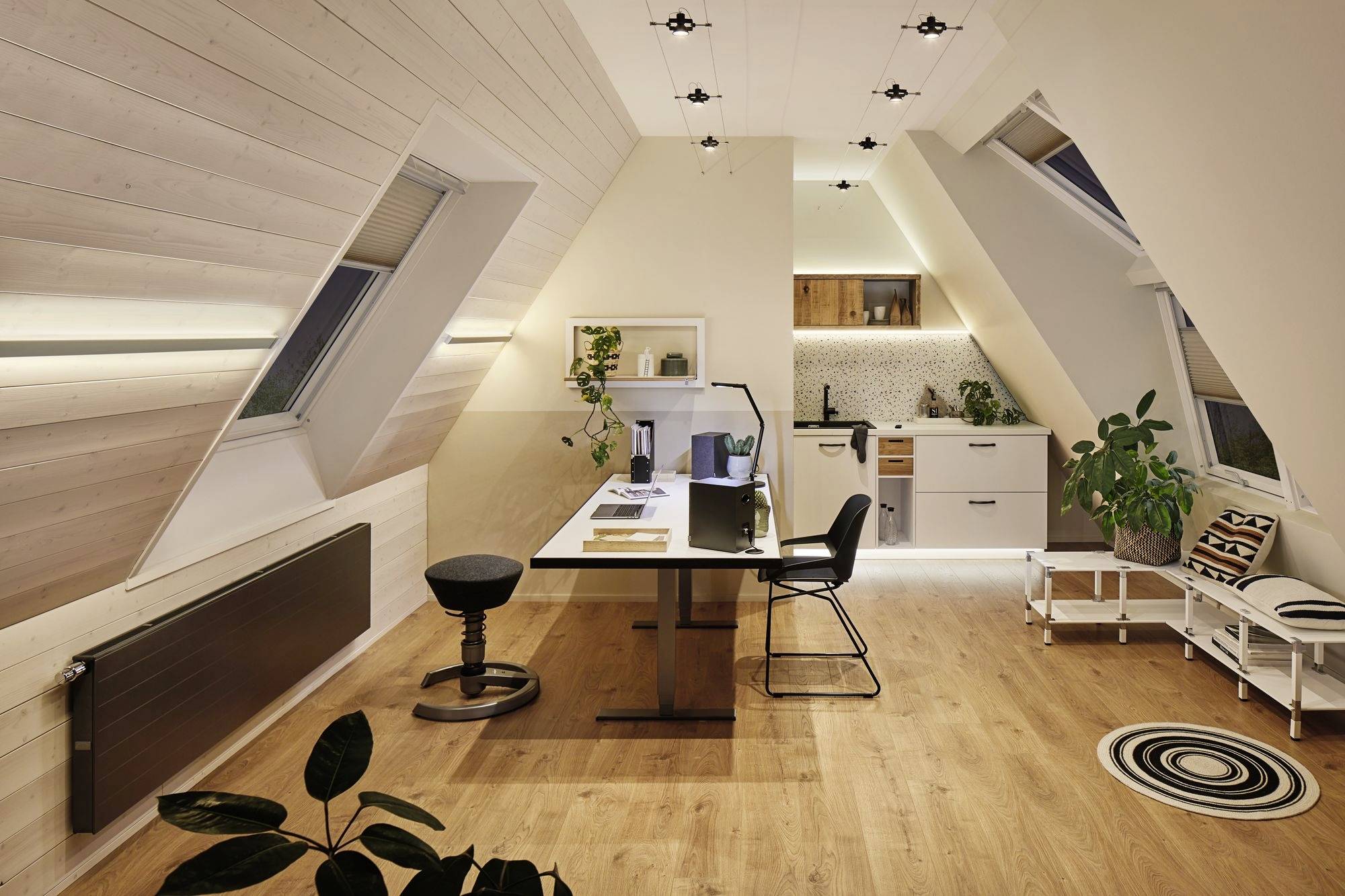 Zolderkantoor met bureau, laptop, stoel en krukken. Moderne geïntegreerde keuken op de achtergrond. Lichte houten betimmering en verlichting.