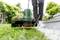 Een persoon gebruikt een groene grastrimmermachine om het gras langs een bestratingsrand te knippen. De omgeving is een verzorgde tuin.