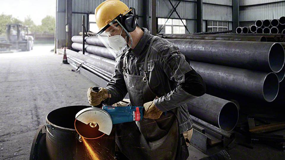 Een arbeider snijdt een metalen buis door met een haakse slijper in een fabriekshall. Hij draagt beschermende kleding, inclusief helm en masker.